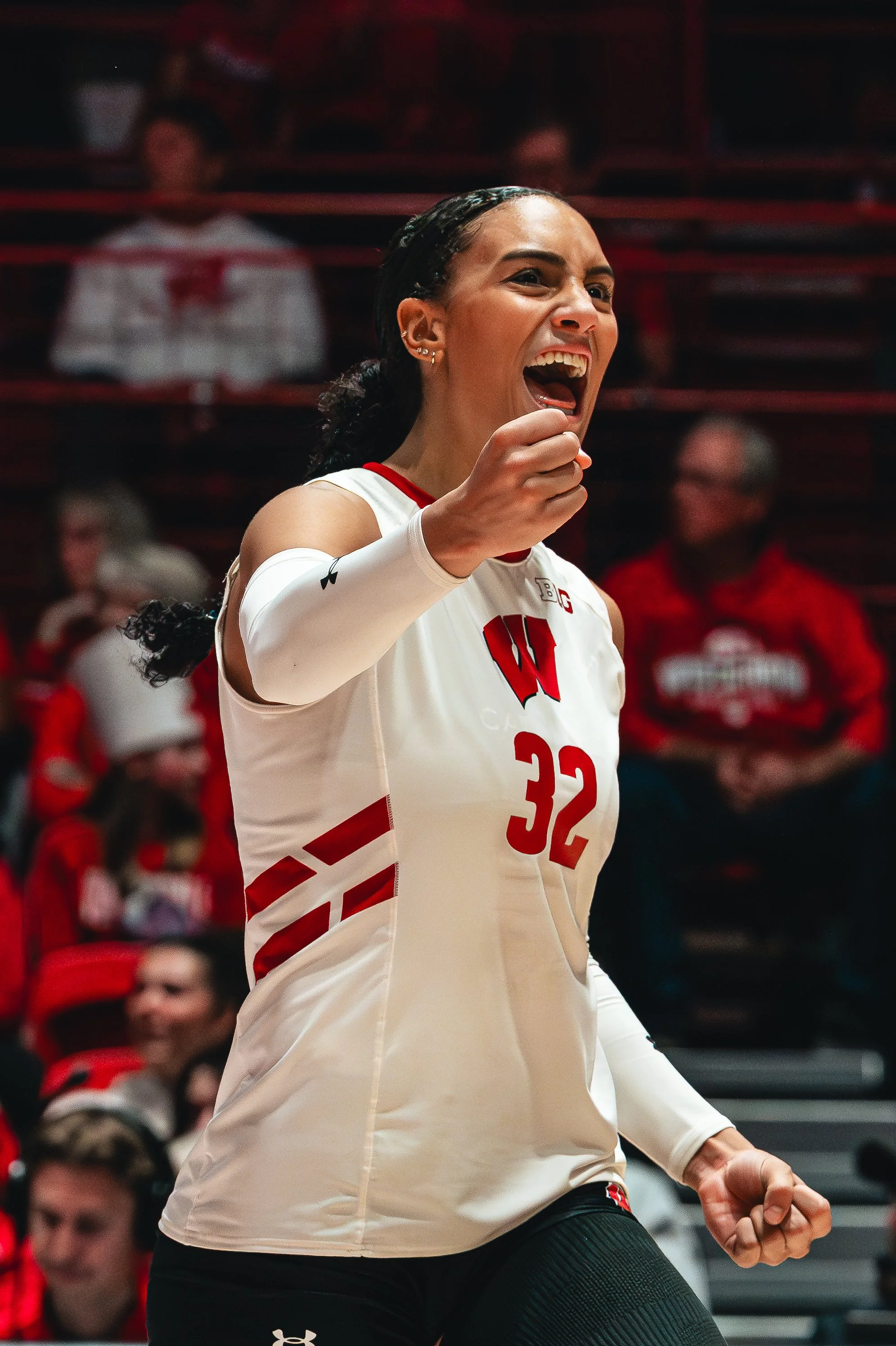 A female volleyball player celebrating on the court in a white jersey with the number 32, with spectators in the background.