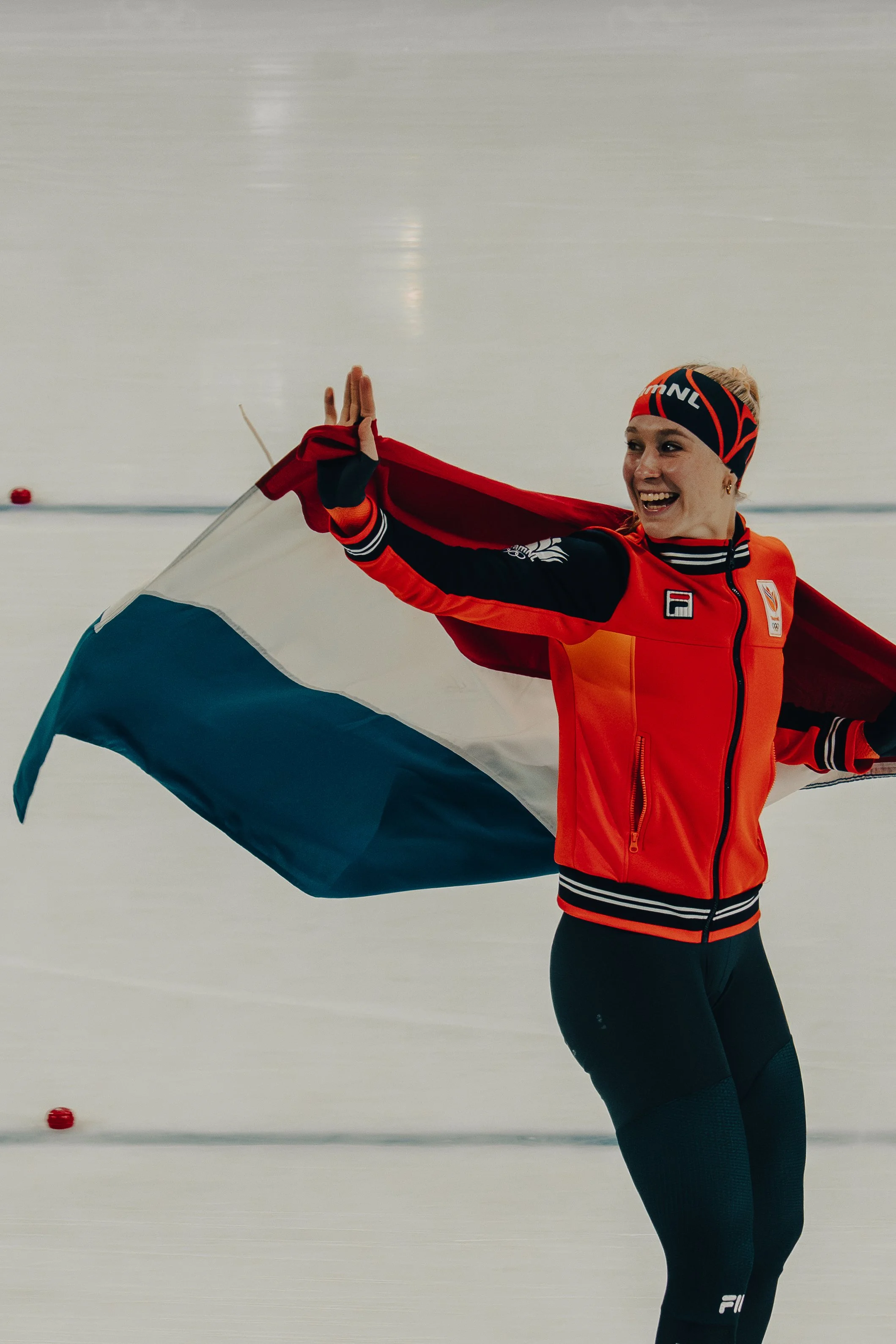 An athlete on an ice rink holding a flag with red, white, and blue colors, smiling and raising her hand in a gesture of victory.