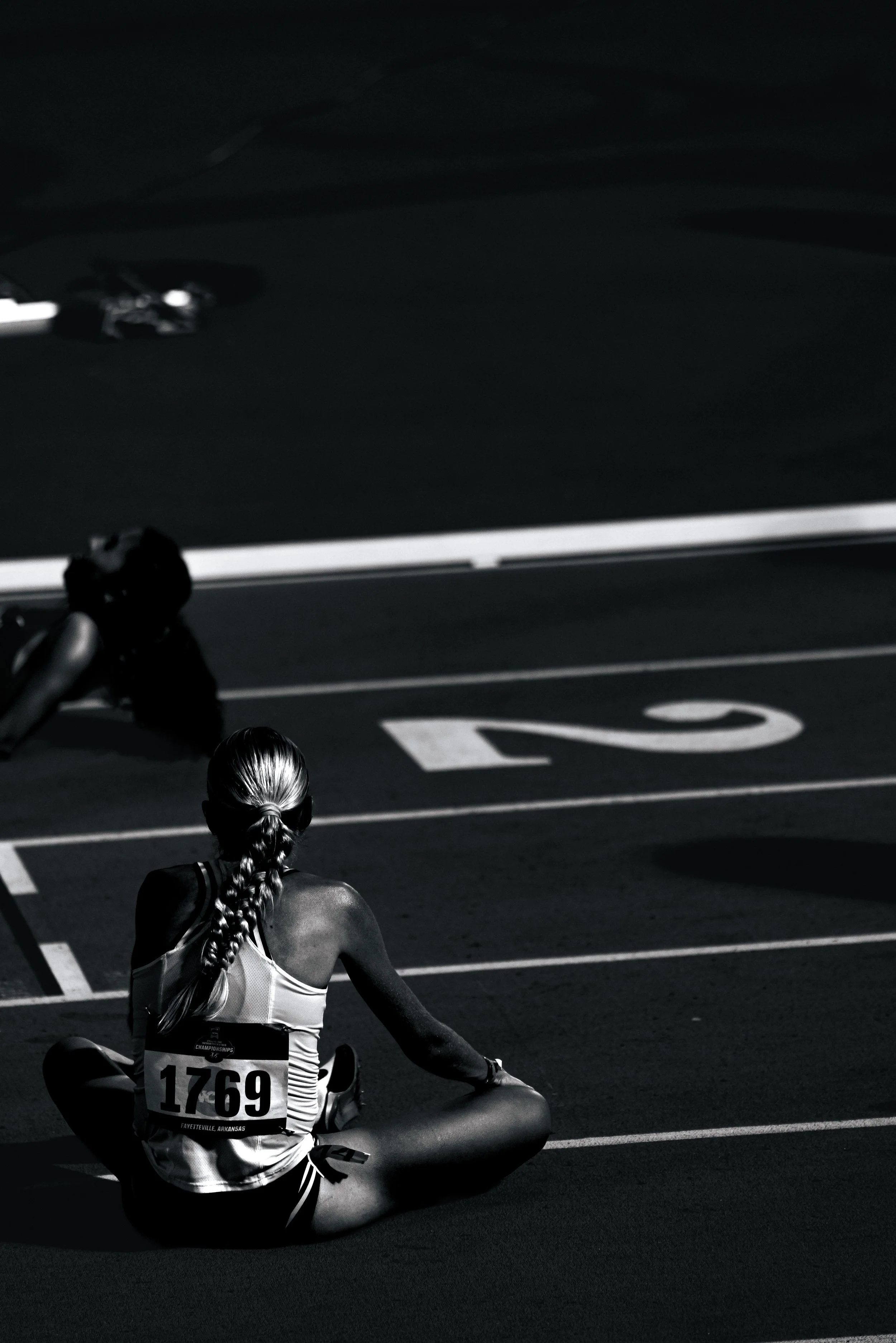 A female athlete with braided hair sitting on the track, wearing sports attire and a bib number 1769, with the words Fayetteville, Arkansas. She is preparing for a race on an outdoor track, with lane markings and a runner in the background.