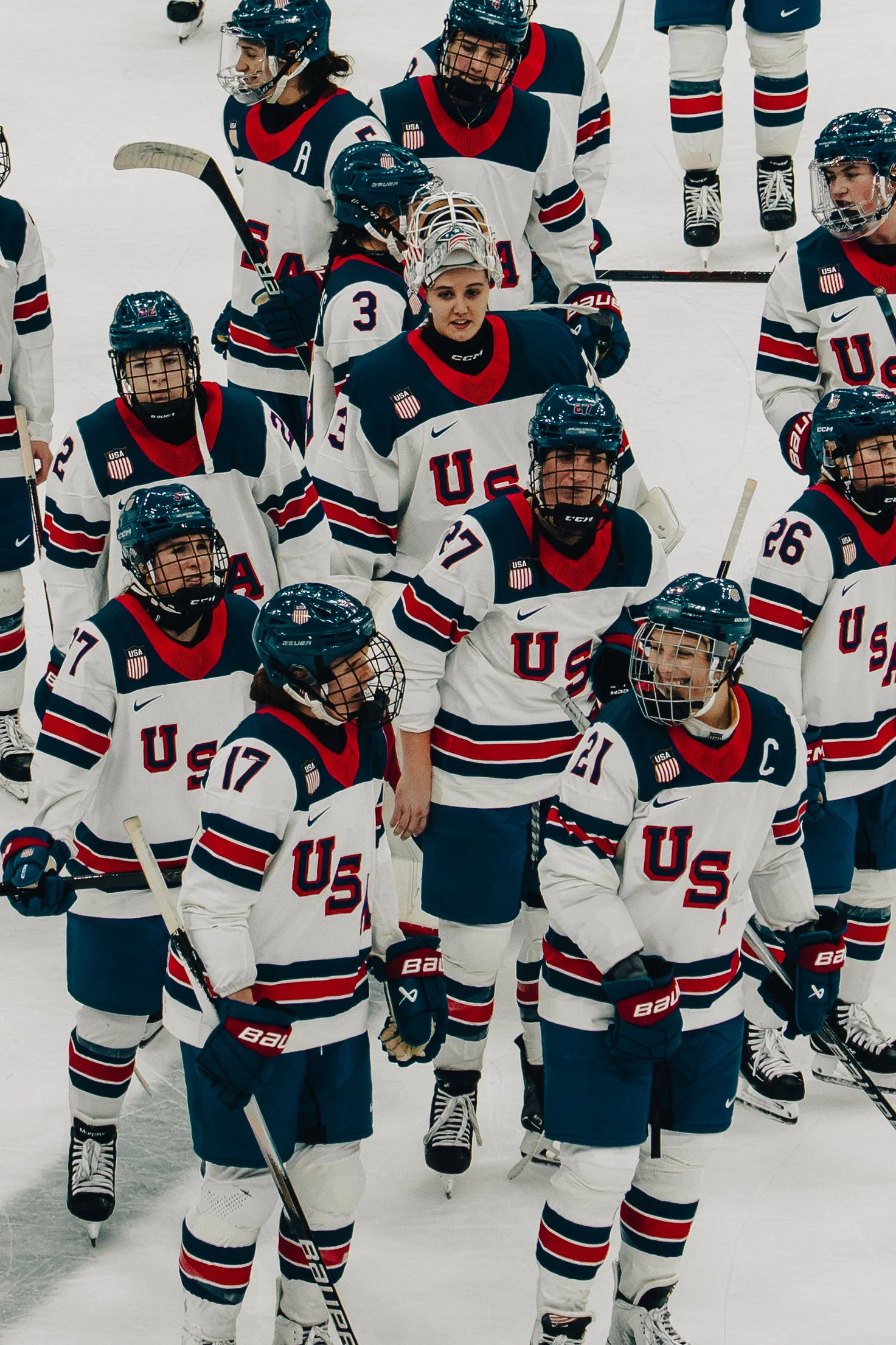 Women’s national ice hockey team on the ice, wearing white jerseys with 'USA' and stars, black helmets, and holding hockey sticks, during a game or practice.