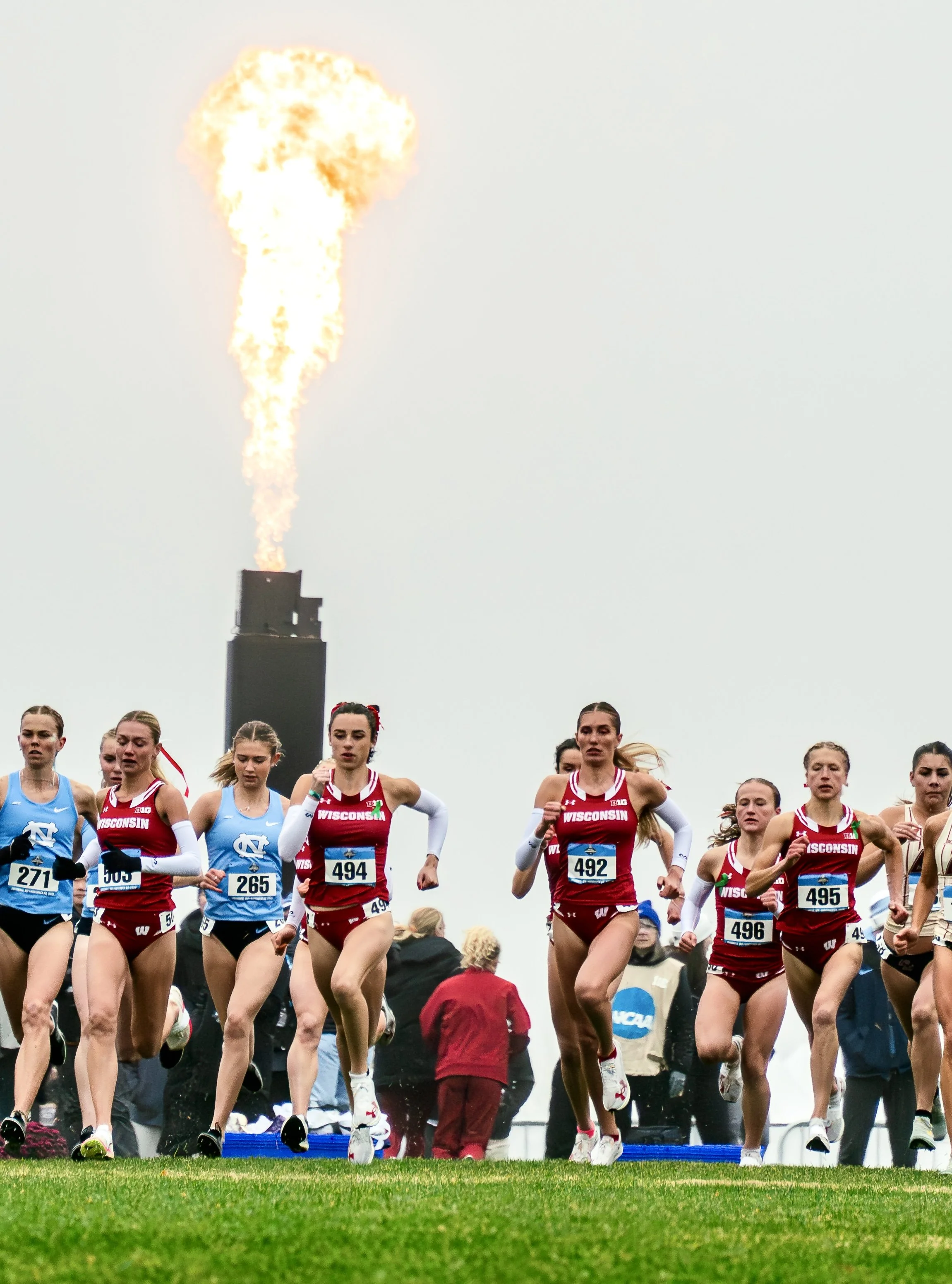 A group of female runners participating in a race, wearing uniforms with 'Wisconsin' and 'NCAA' logos, running on a grassy field with a group of spectators and officials in the background. The sky is overcast, and there is a large industrial smokesta