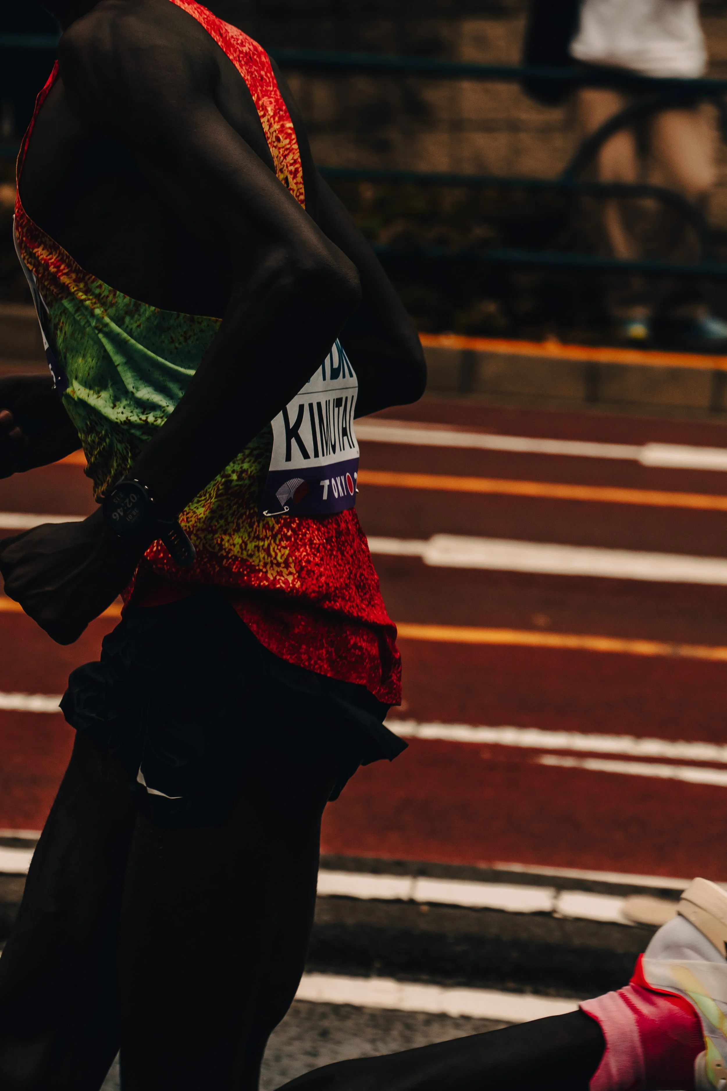 Runner wearing a colorful shirt and black shorts on a track at night, with others running in the background.
