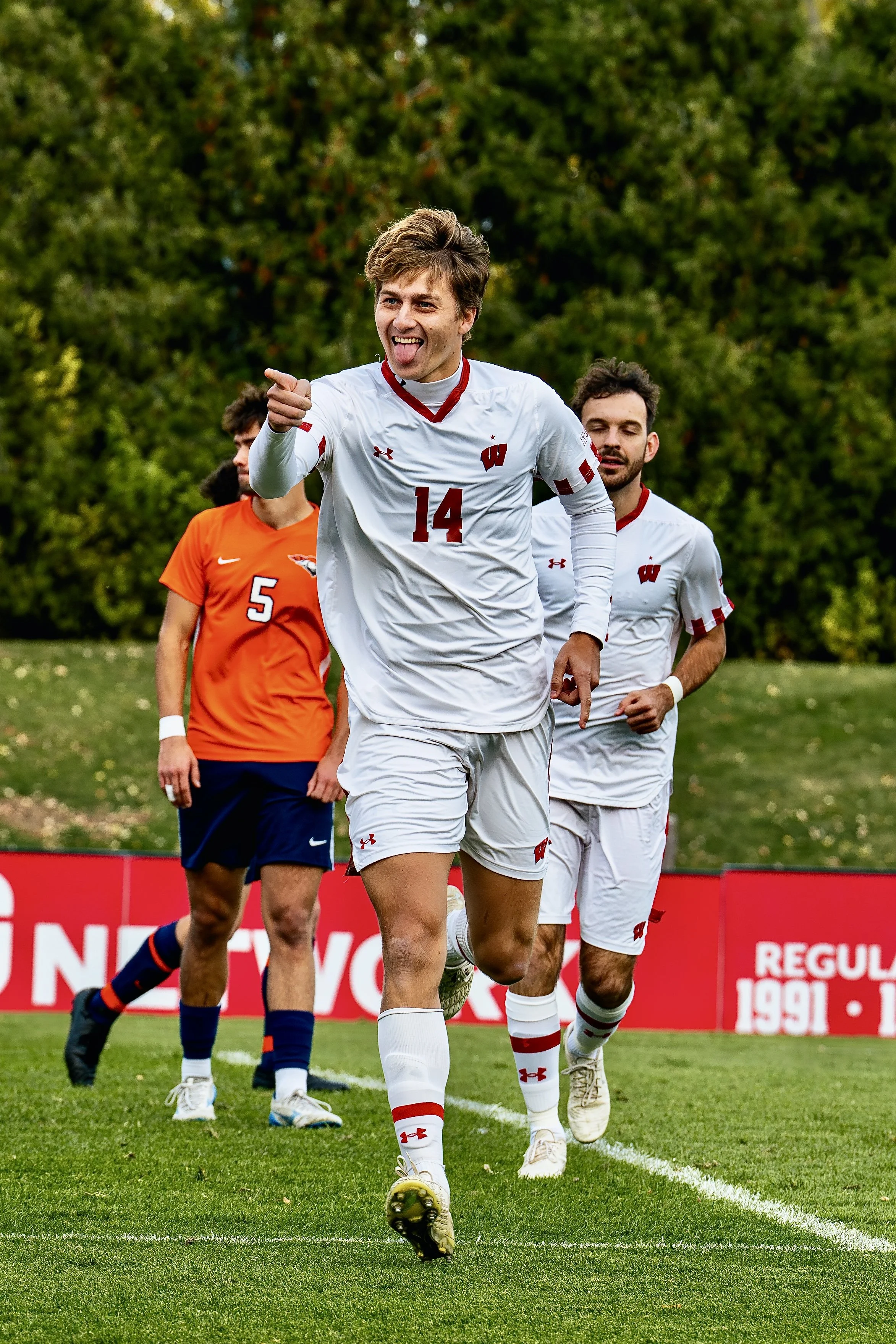 Soccer players running on the field, the player in front is wearing a white uniform with number 14 and is pointing forward, others in background wear orange and white uniforms, trees in background.