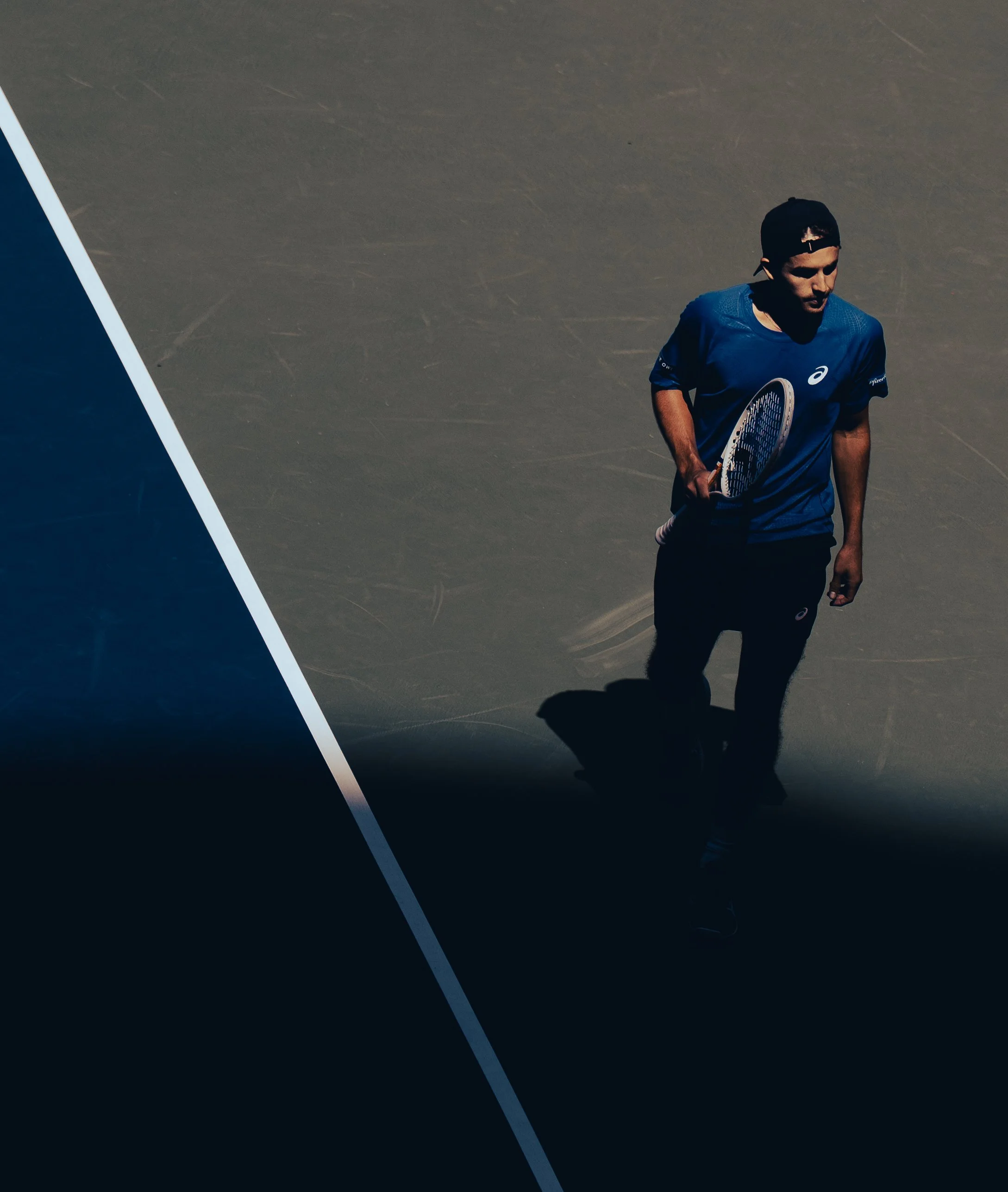 A male tennis player in a blue shirt and black cap holding a tennis racket on a court with a faded green surface.