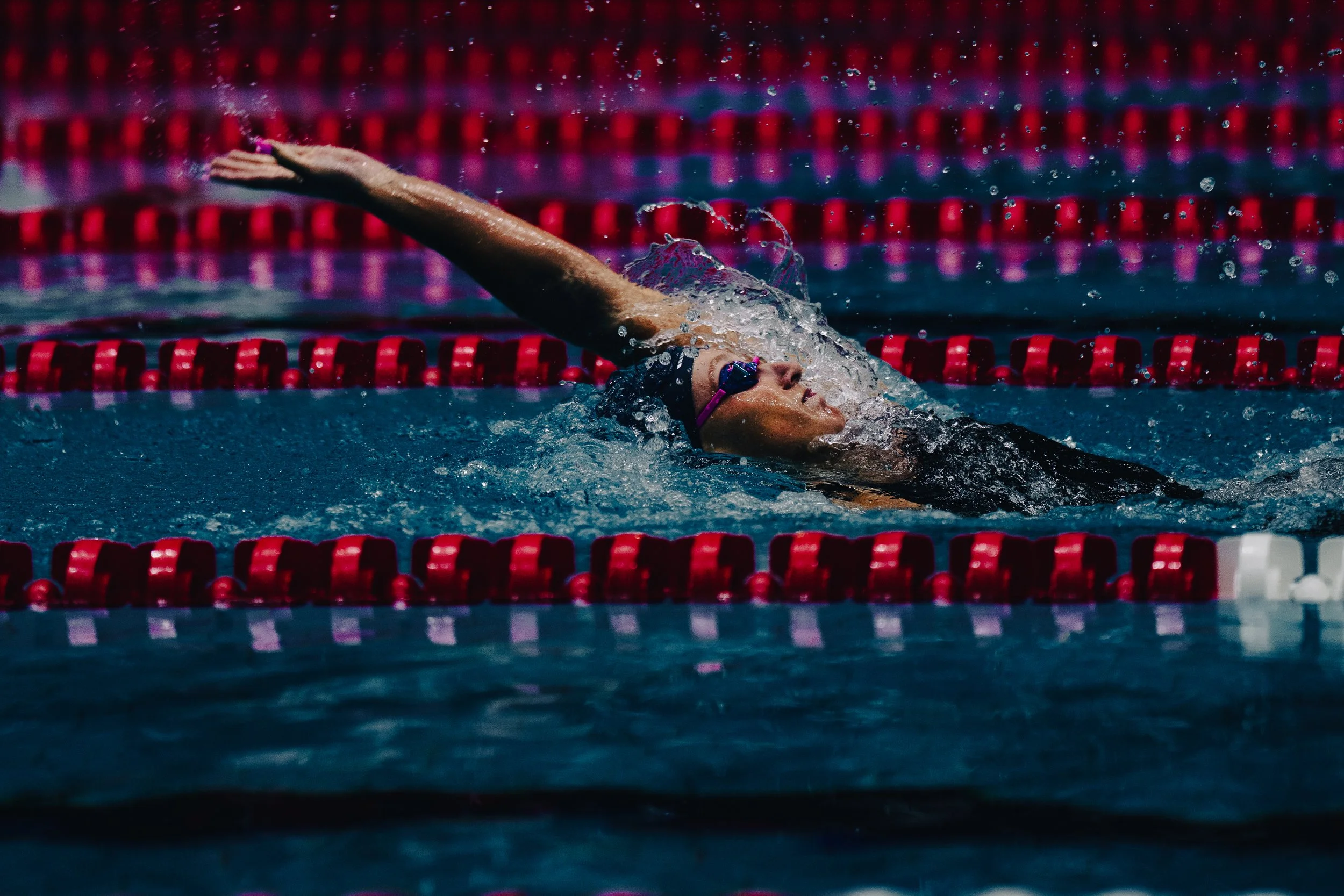 Swimmer in a competitive swimming pool performing a stroke, wearing goggles and a swim cap, with lane dividers visible.