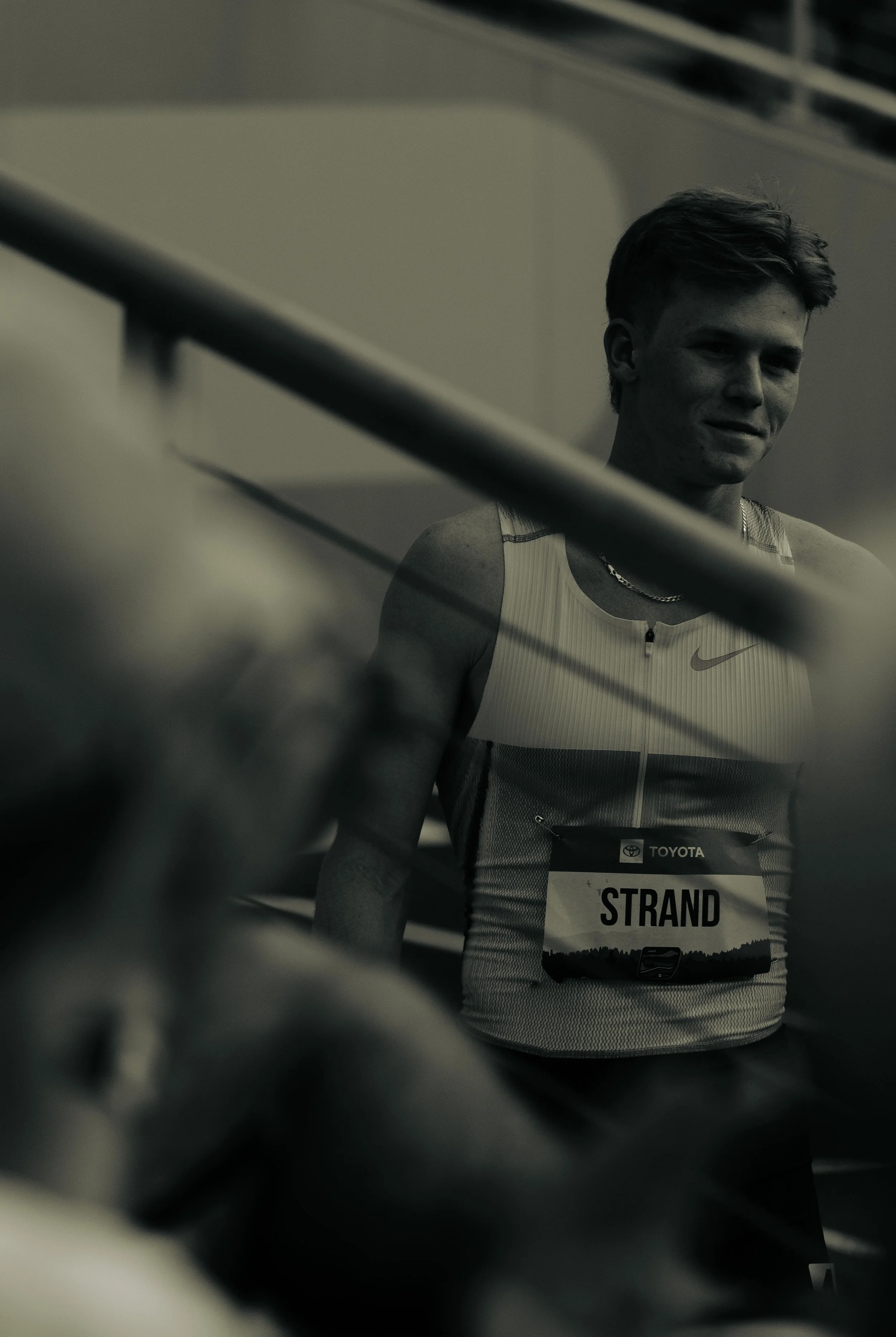A male athlete with short dark hair, wearing a sleeveless athletic shirt with the Nike logo, looking at the camera. He is wearing a bib with the name 'Strand' and the number 185, and a chain necklace. The photo appears to be taken during a race or co