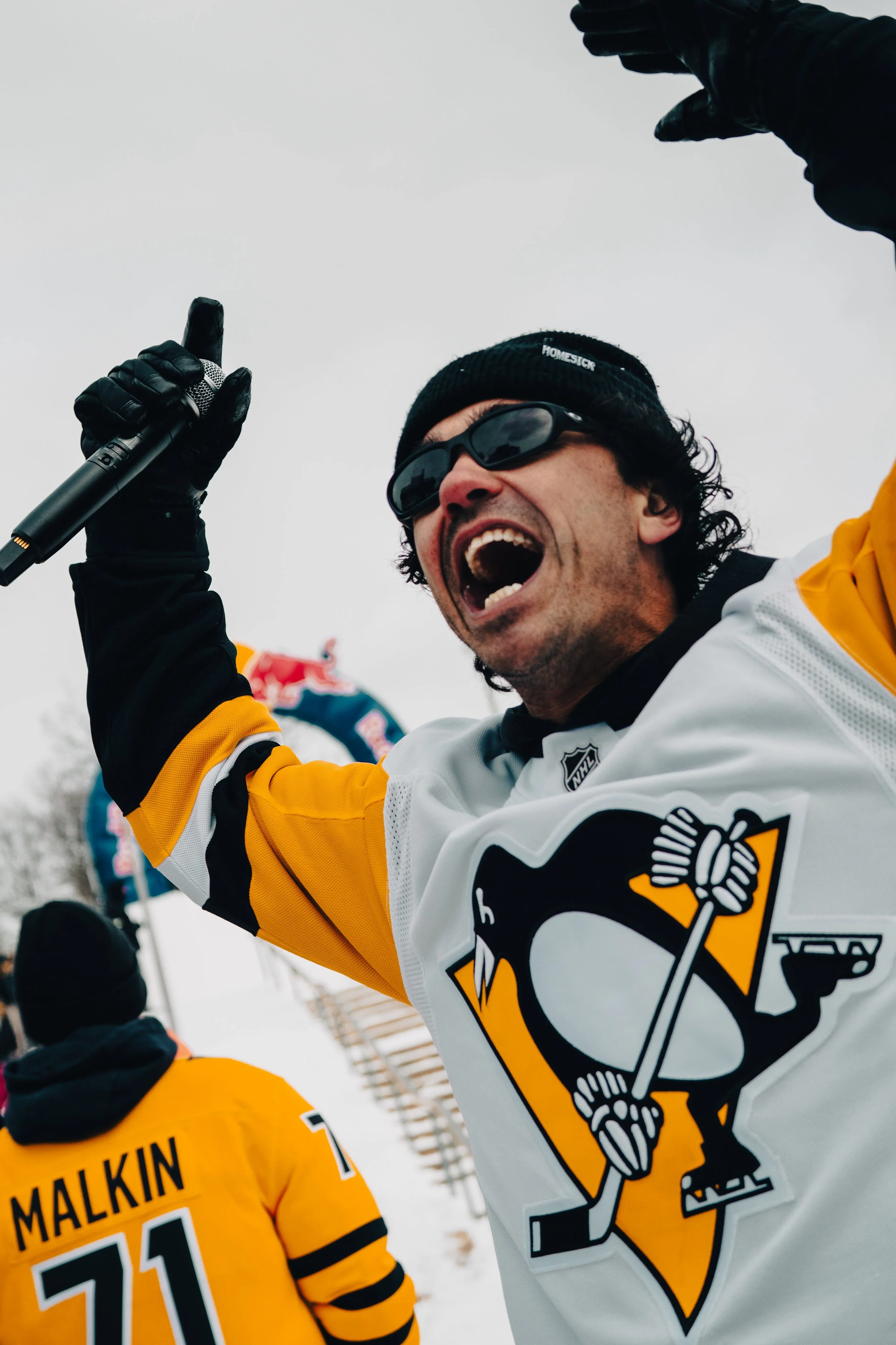 A man wearing black sunglasses, black gloves, a black beanie, and a white and yellow hockey jersey celebrates outdoors in a snowy environment, holding a microphone and raising his right hand.