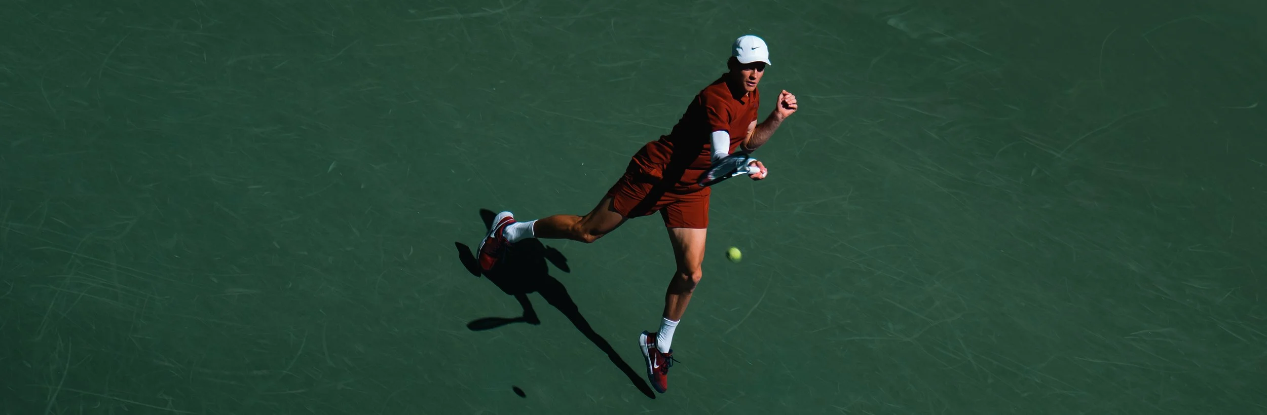 A male tennis player wearing a white cap, orange shirt, orange shorts, and red shoes running on a green tennis court while holding a tennis racket, with a tennis ball in the air near him.