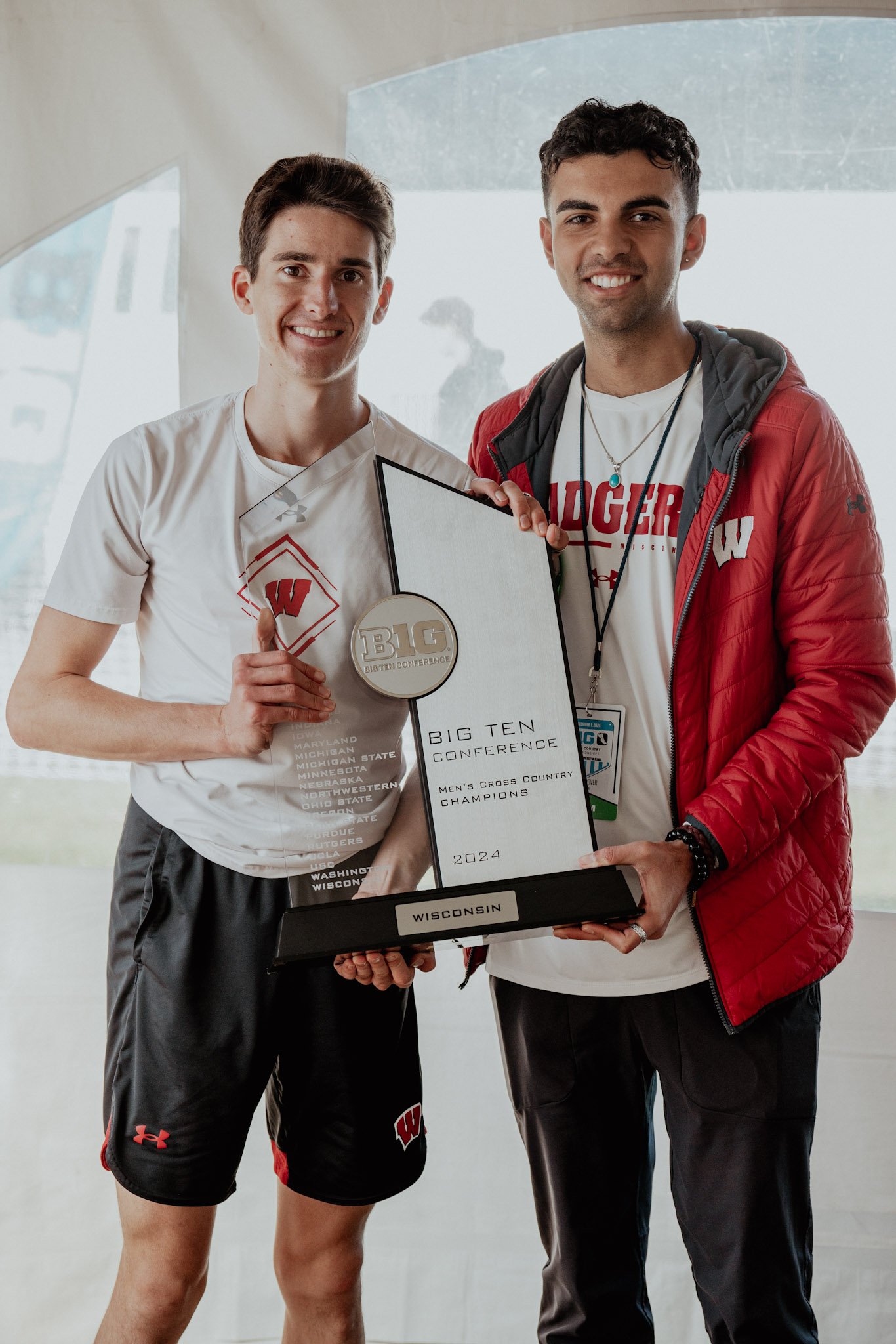 Two young men smiling and holding a trophy and certificate for Wisconsin men's cross country champions at the Big Ten Conference in 2024. They are indoors, one wearing a white frame shirt and black shorts, the other in a white shirt and red jacket.