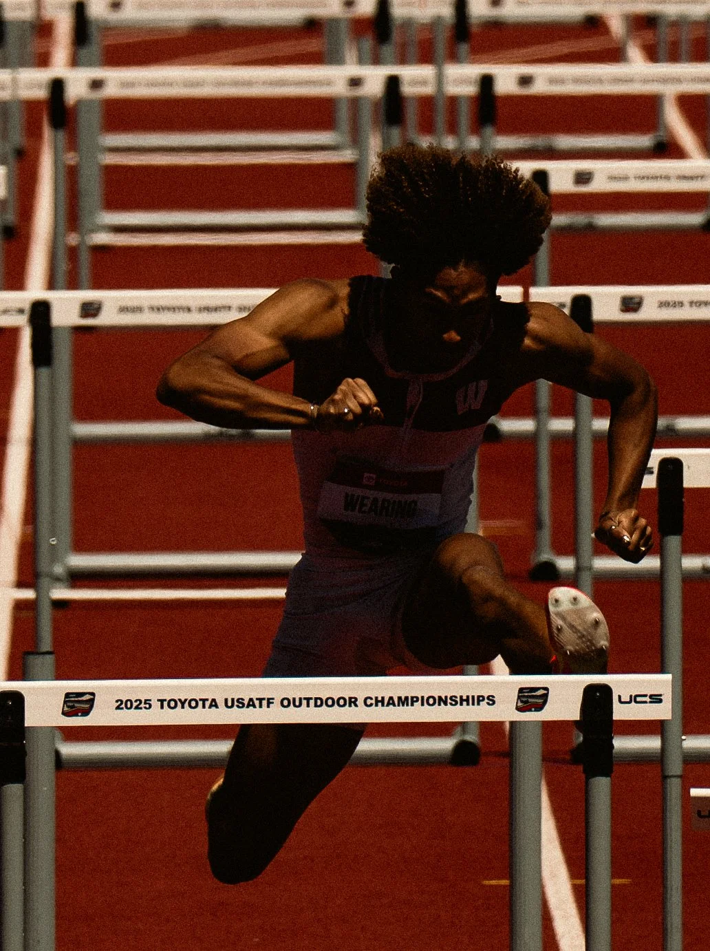 An athlete with an afro hairstyle clearing hurdles during a race at the 2025 Toyota USATF Outdoor Championships, wearing a black and purple USA team uniform on a red track.