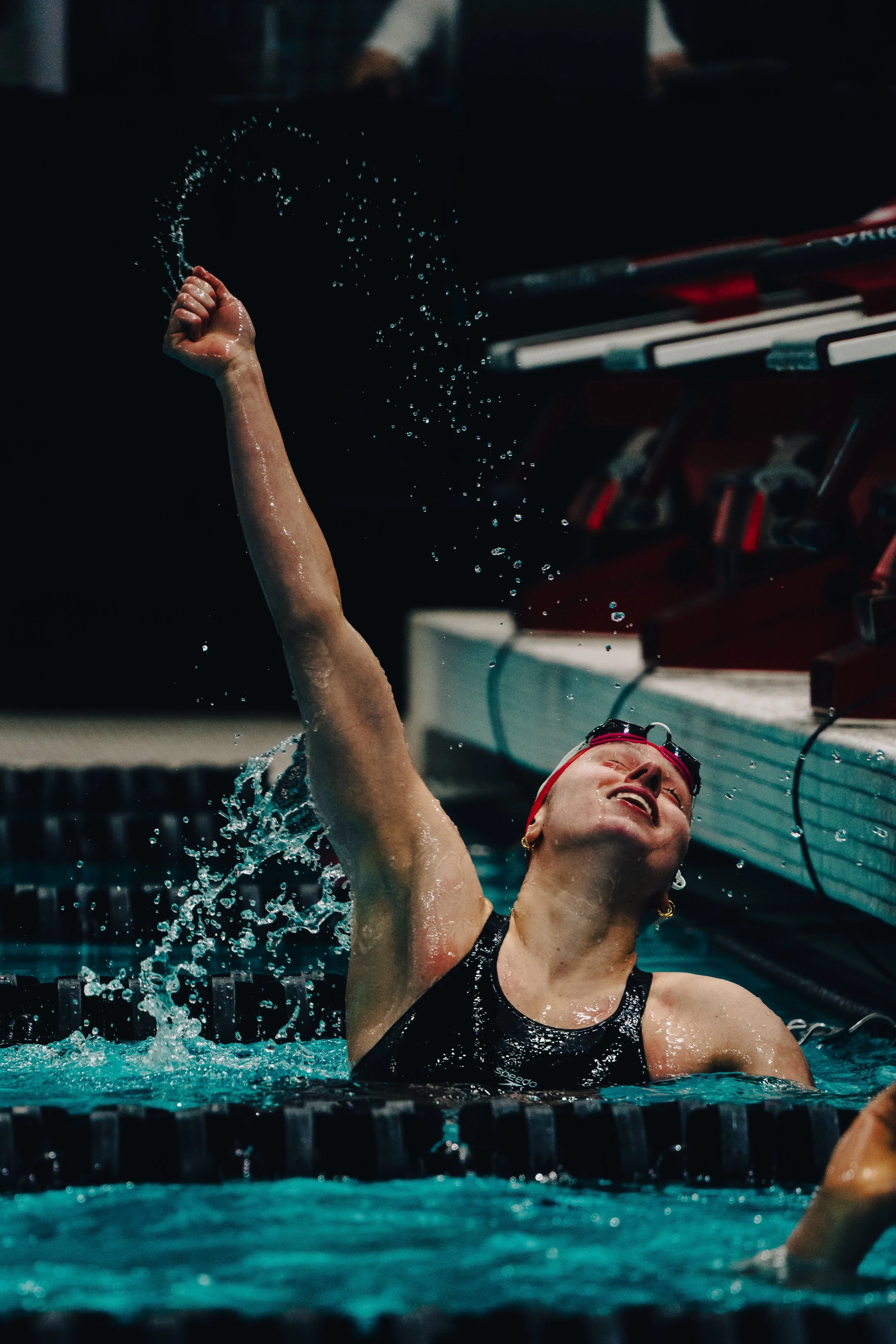 A female swimmer in a black swimsuit and red swim cap, with goggles on her forehead, raising her arm out of the water in a swimming pool, celebrating or finishing a race, with water splashing around her.