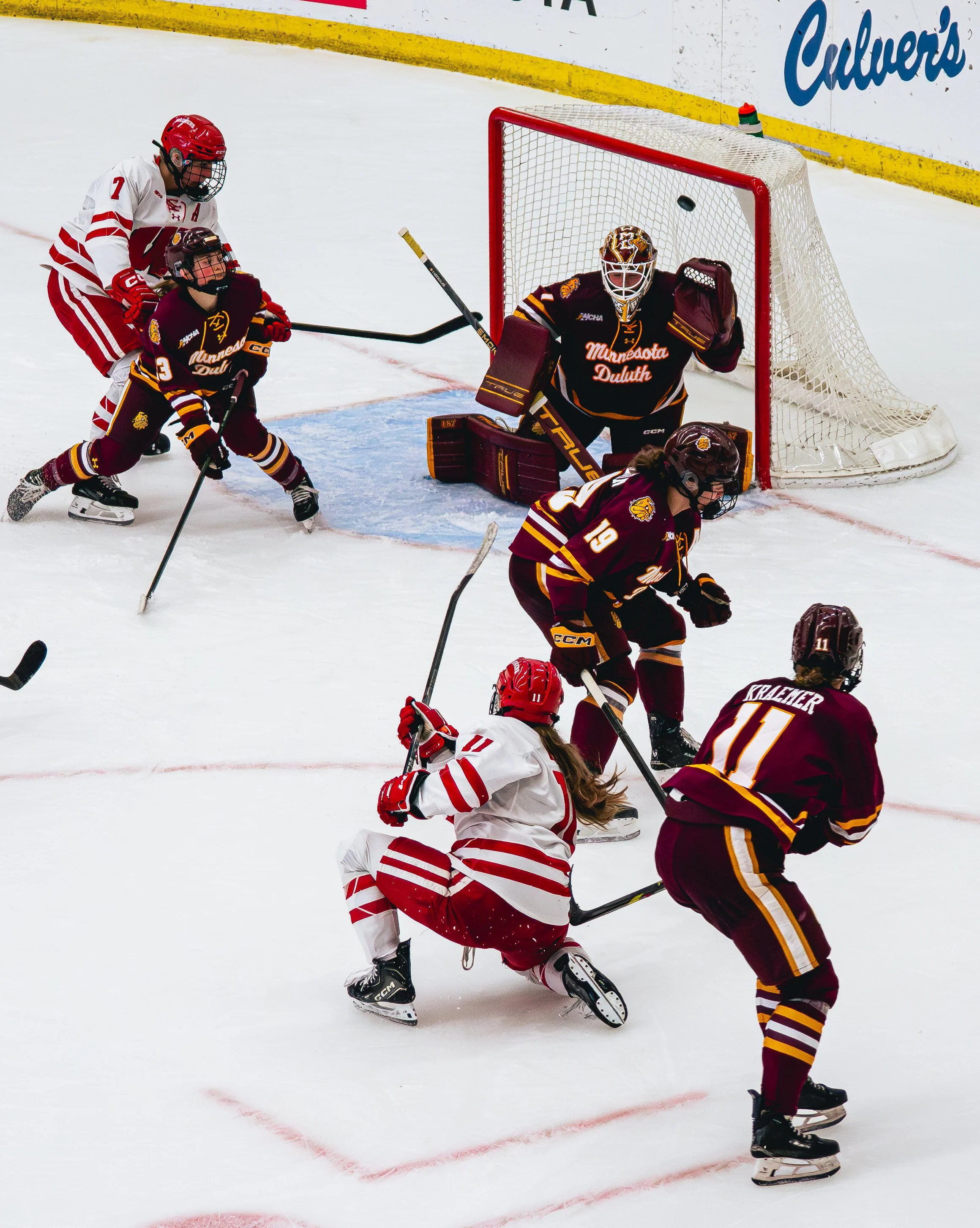 A hockey game with players from two teams competing near the goal, with one player in a white and red uniform falling on the ice and a skater in a maroon and gold uniform approaching, while the goalie in maroon and gold guards the net.