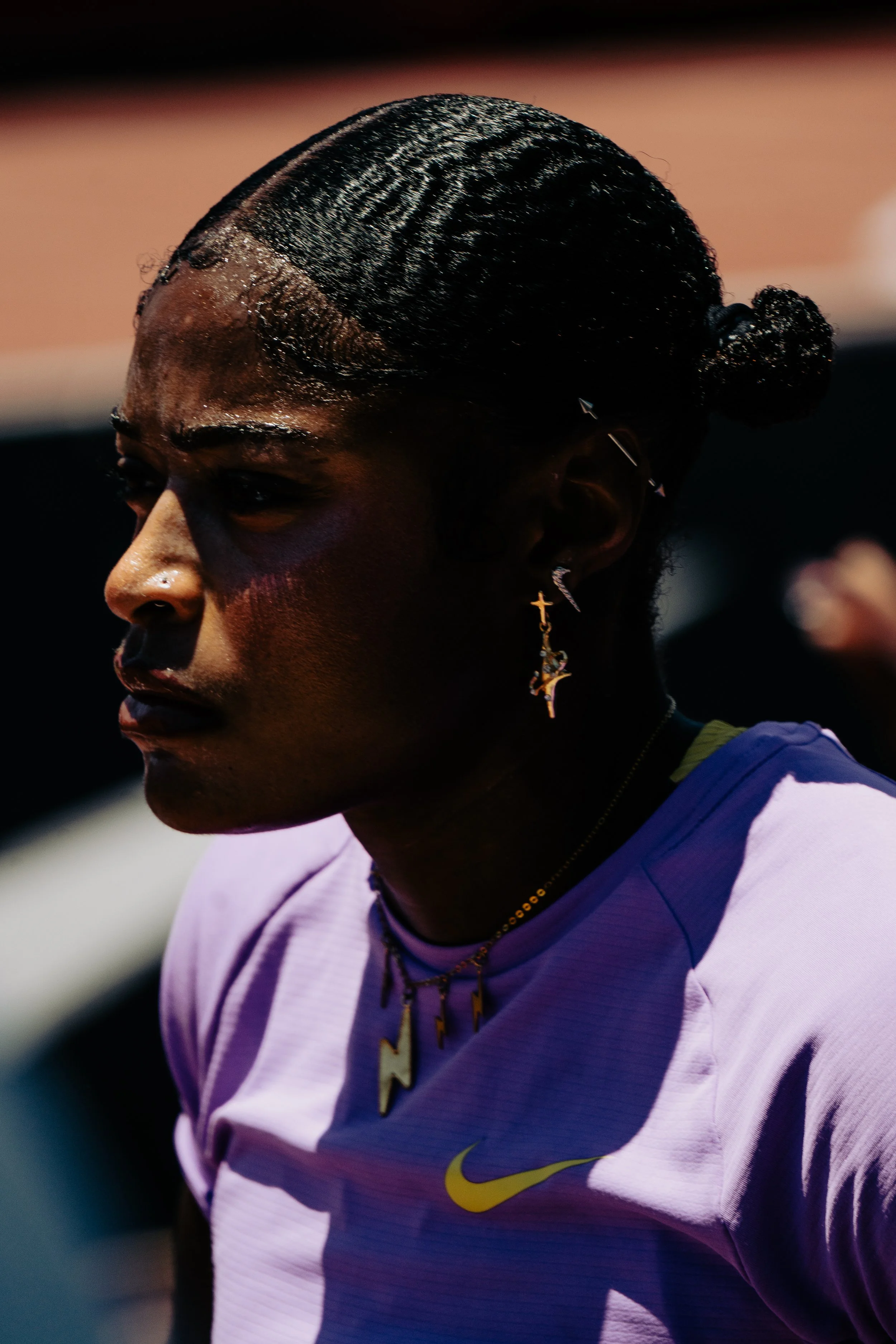 Close-up of a woman with sleek, dark hair tied in a bun, wearing earrings and a purple sports shirt with yellow accents, in an outdoor setting.