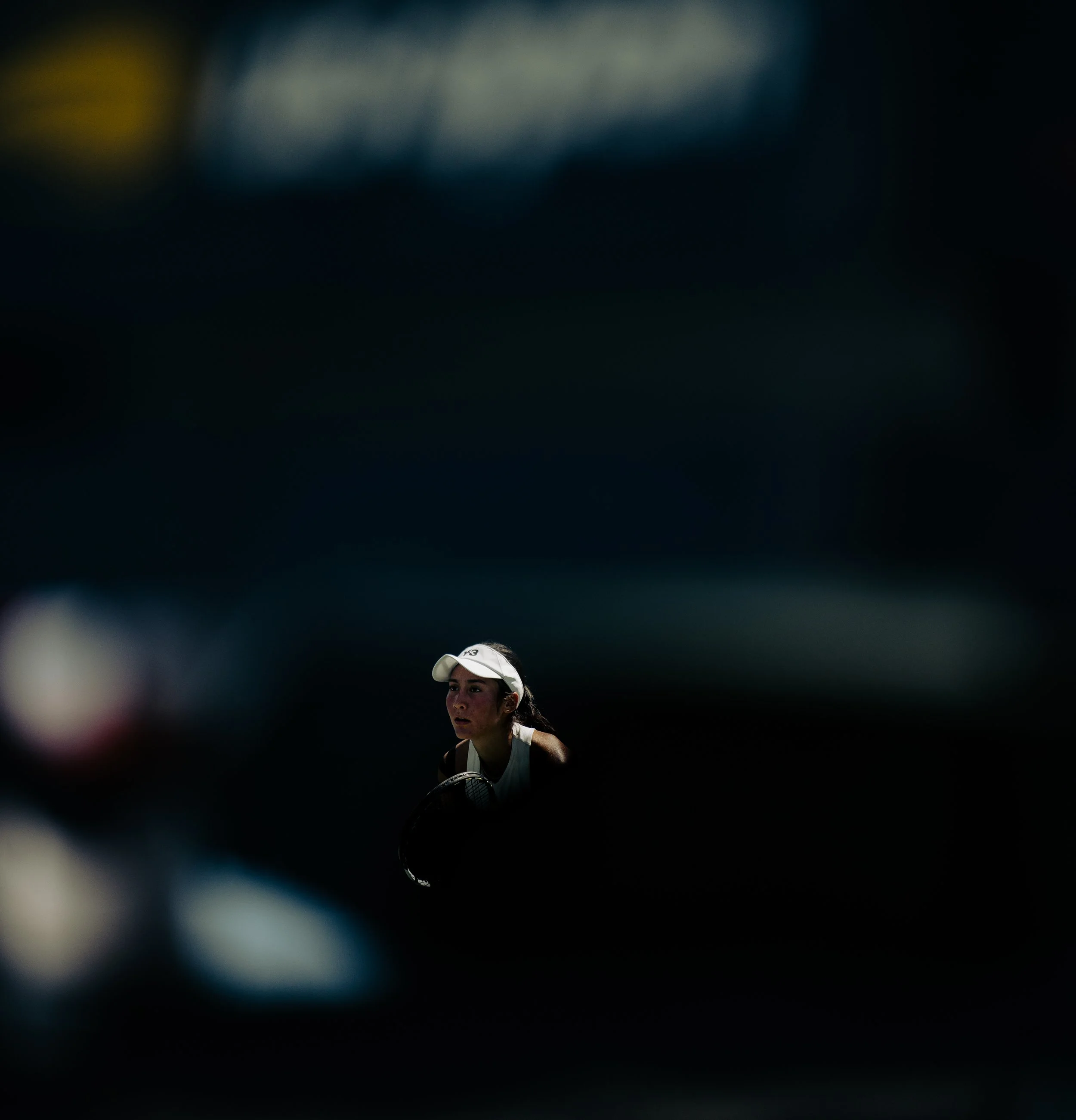 A female tennis player wearing a white cap and white athletic outfit, holding a tennis racquet, in a dark background.