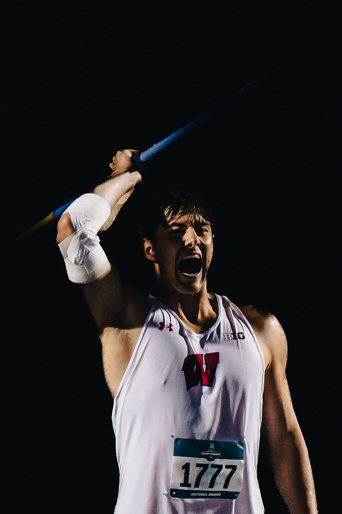 A male athlete in a white jersey with the number 1777, celebrating with his mouth open and holding a torch above his head during the Olympic torch relay.