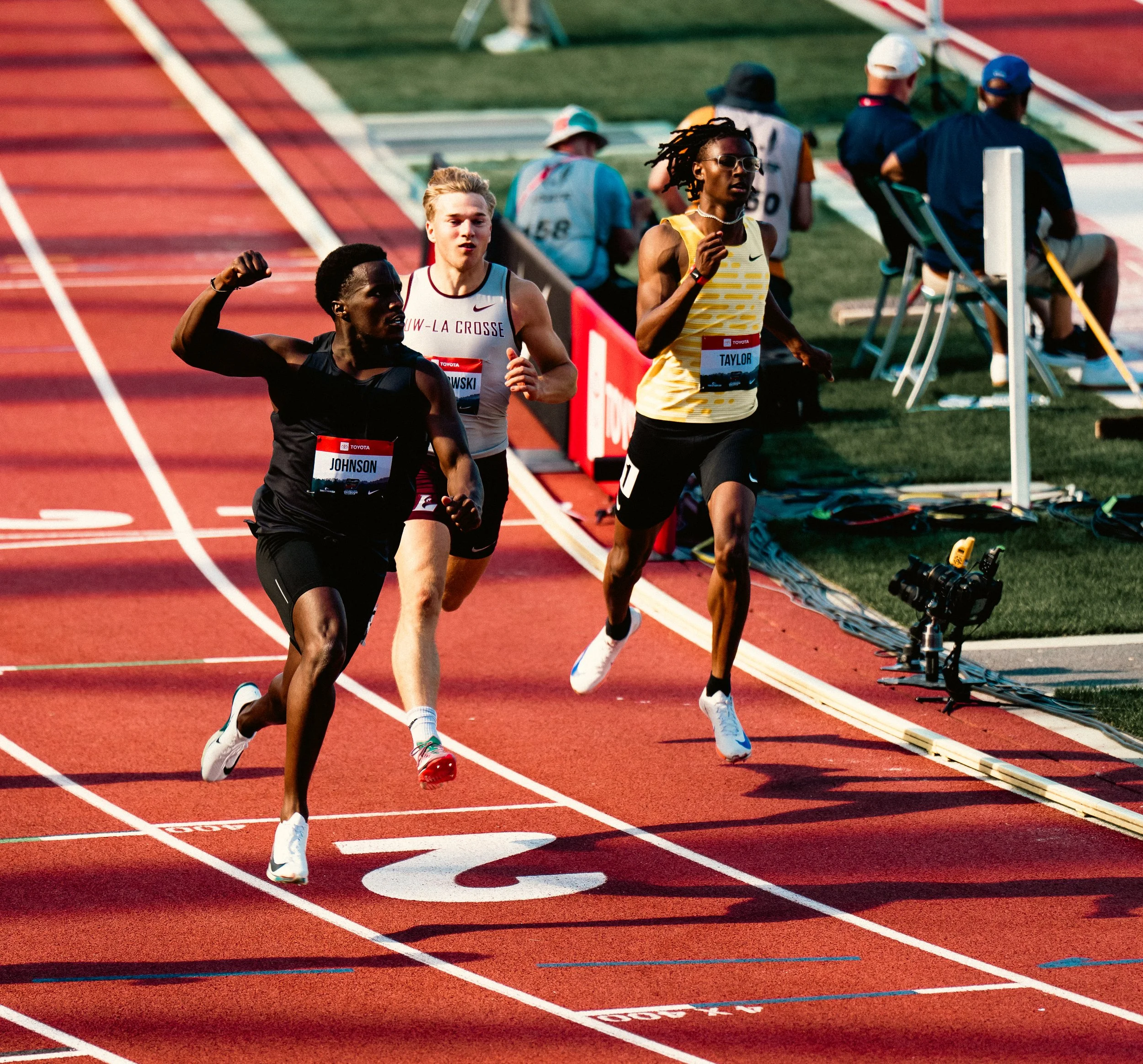 Three athletes competing in a track race, running on a red outdoor track with white lane markings, while officials and spectators watch in the background.