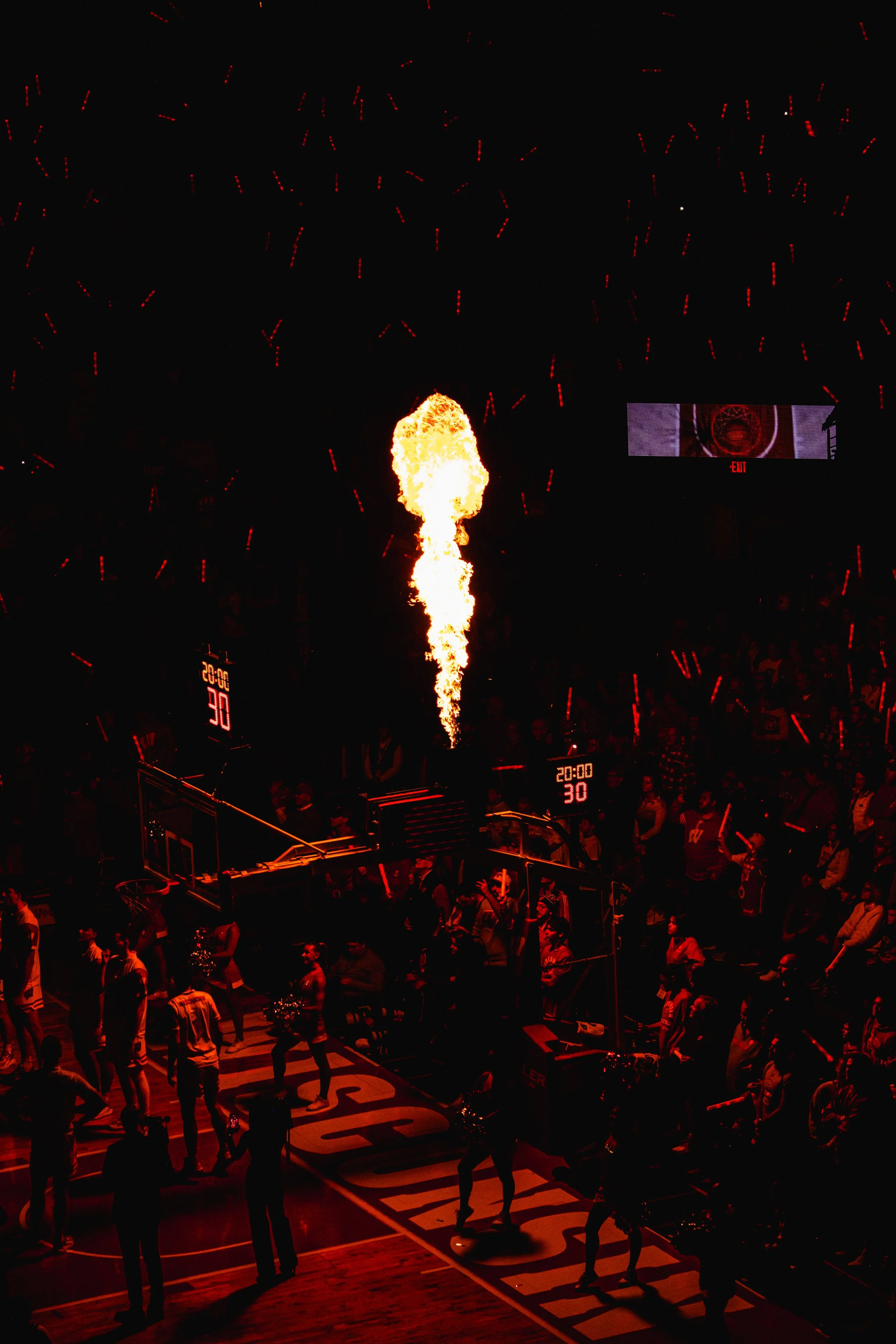 Basketball arena filled with spectators, with a flame on the court and fireworks in the dark sky.