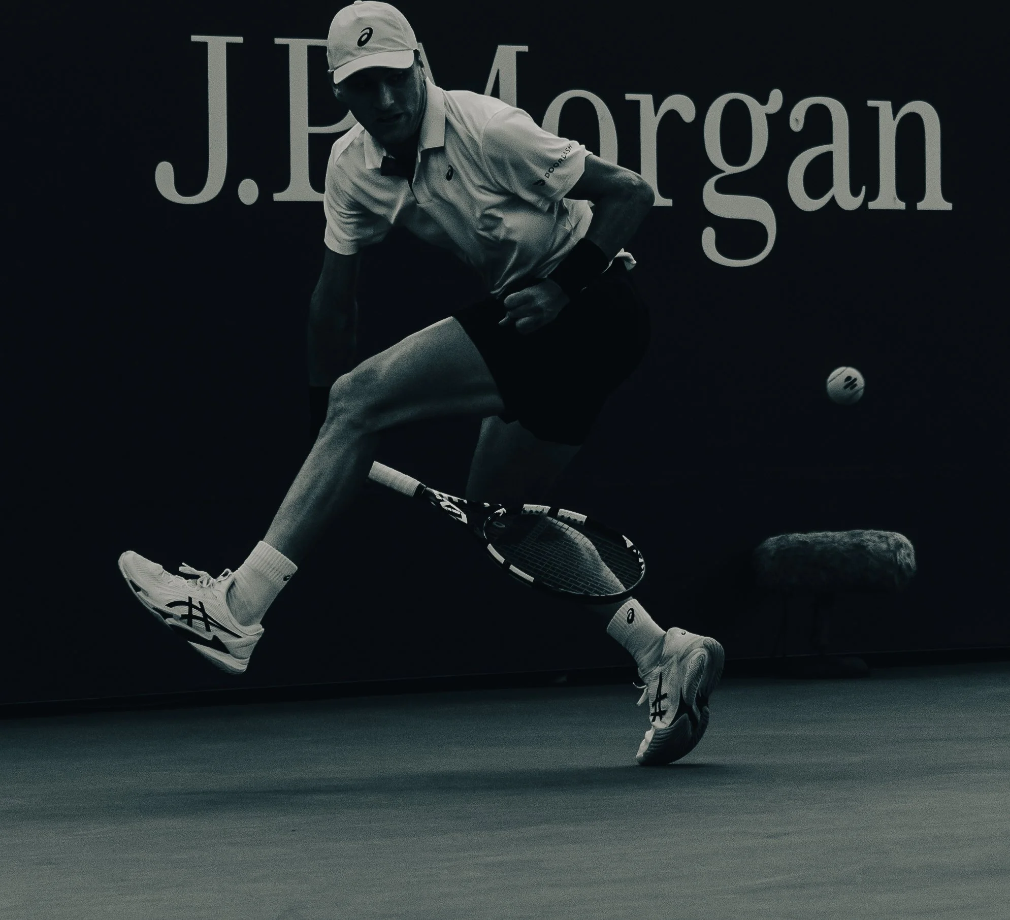 A black-and-white photo of a man in athletic wear playing tennis, wearing a cap and sneakers, with a tennis ball in motion on the court.