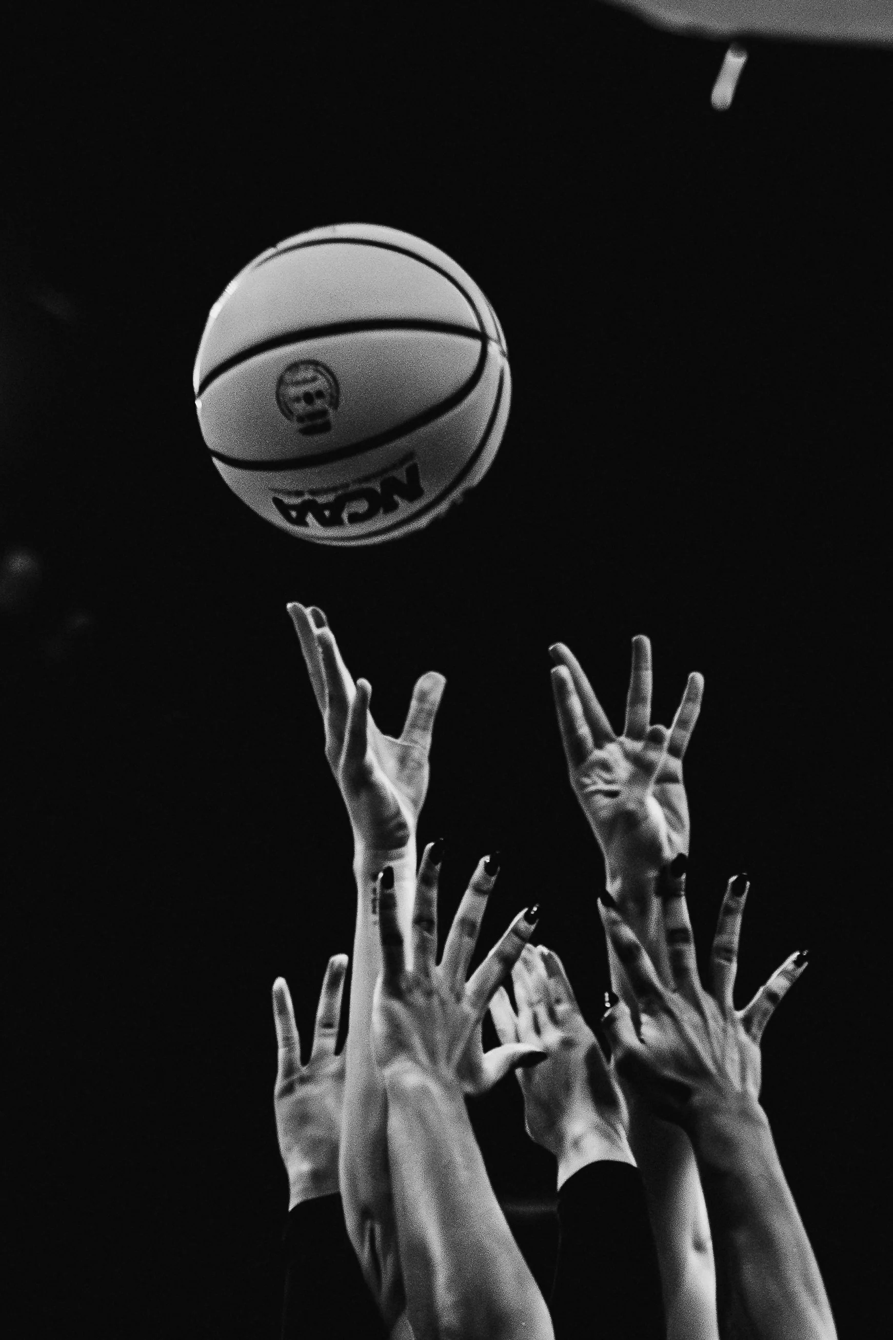 Black and white image of multiple hands reaching up towards a basketball in the air. The basketball is labeled with the NBA logo.