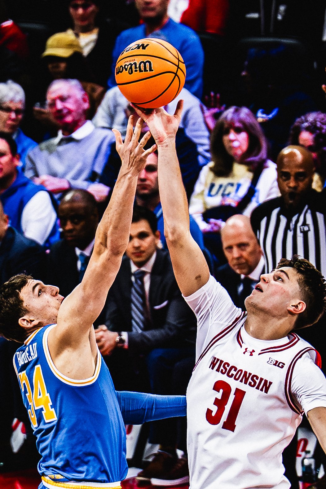 Two basketball players from opposing teams reaching for the ball during a game, with a crowd of spectators in the background.