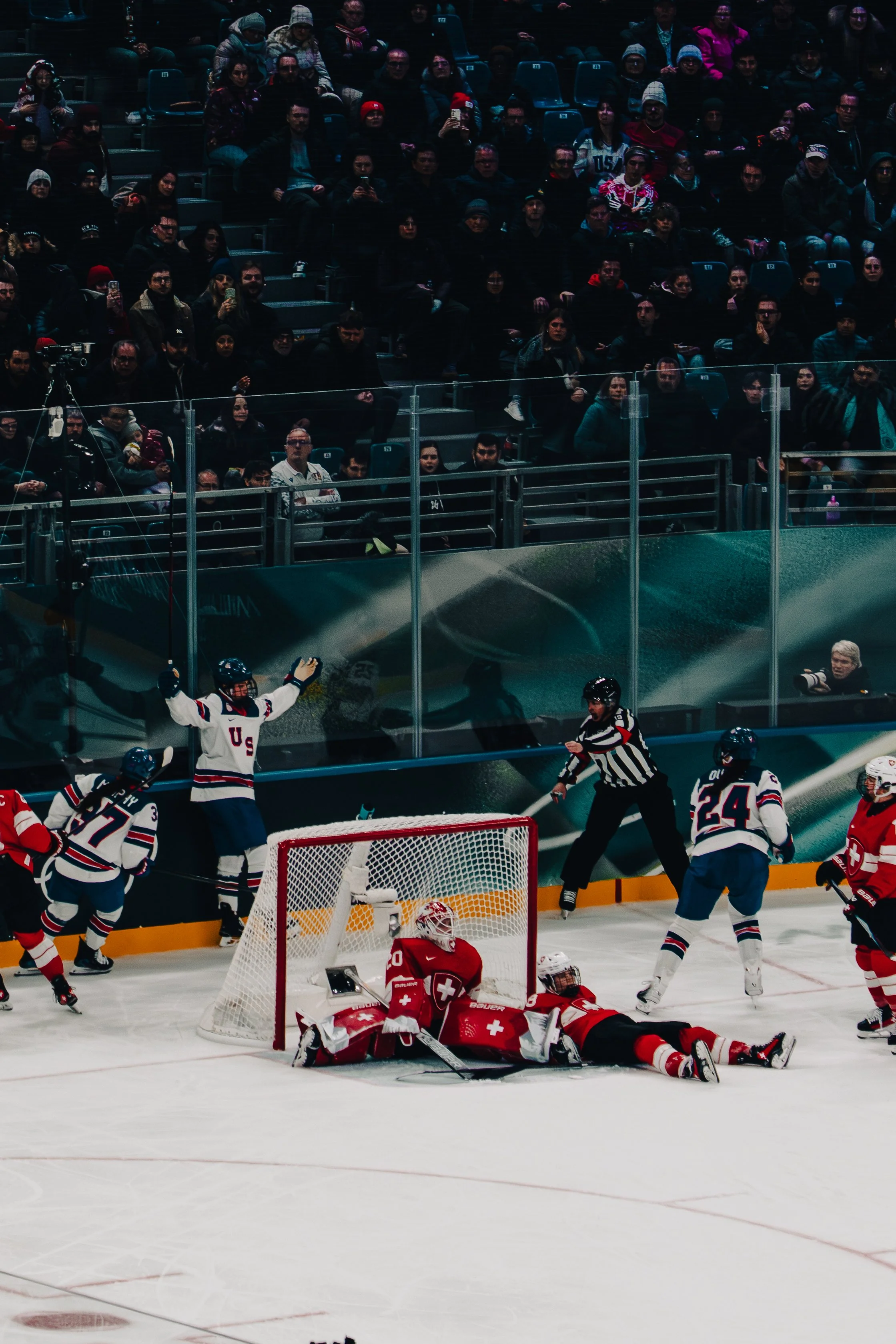 An ice hockey game in progress with players, a referee, and a crowd in the stands. Some players are near the goal, likely celebrating a goal, while the crowd watches intently.