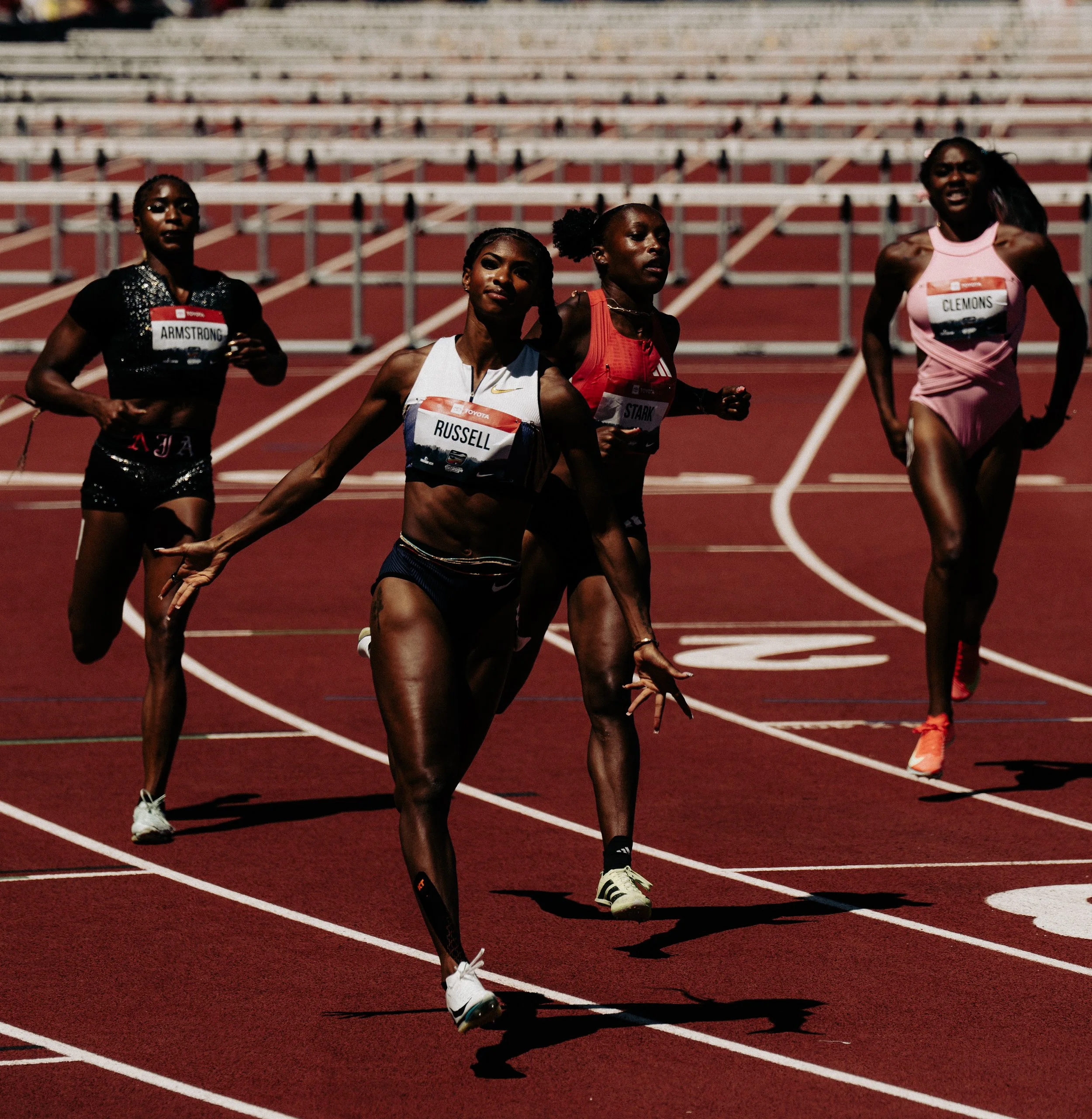 Five female athletes running on a red outdoor track during a race, with empty bleachers in the background.