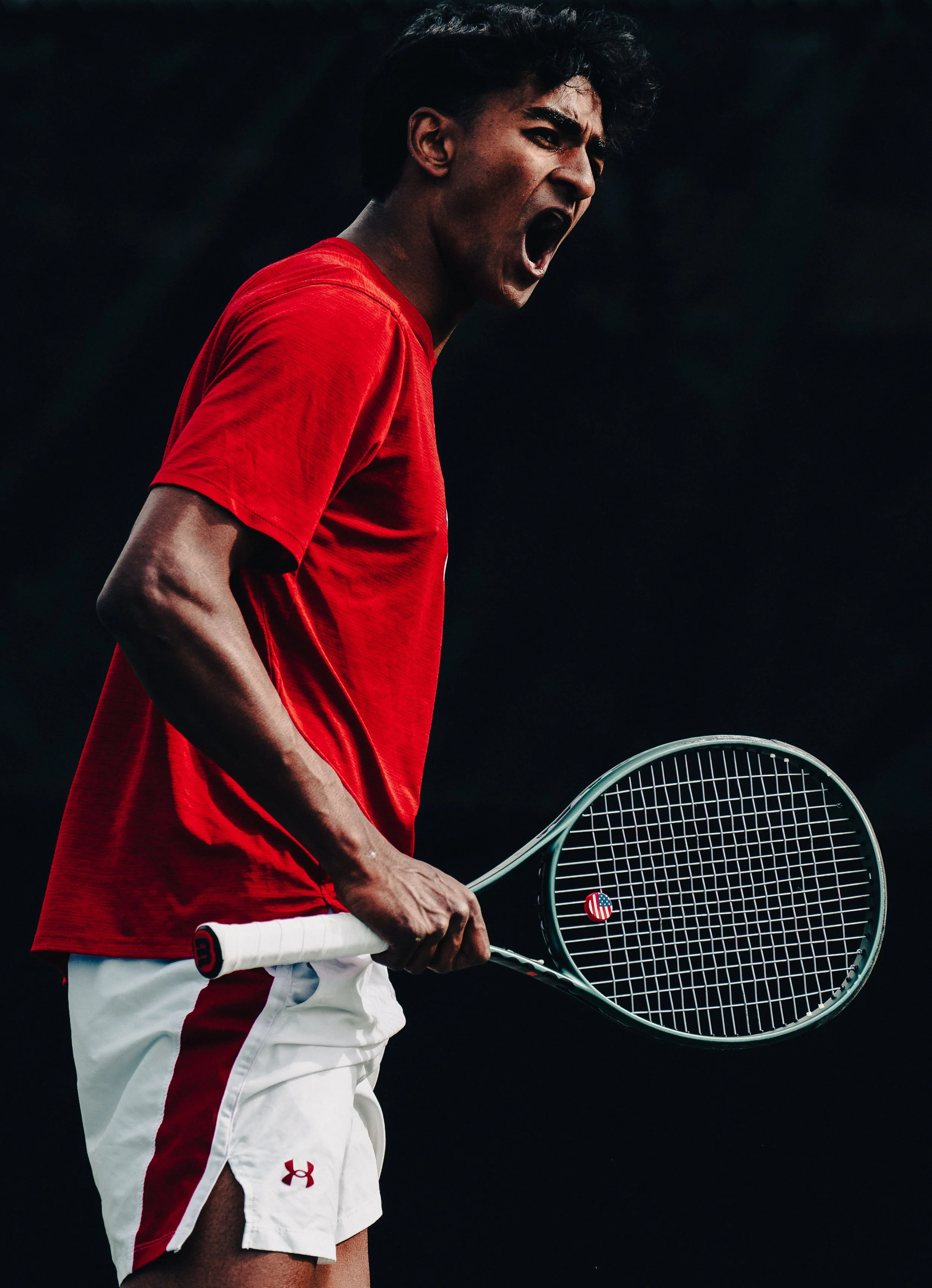 A male tennis player wearing a red shirt and white shorts, holding a tennis racket, shouting on a dark background.