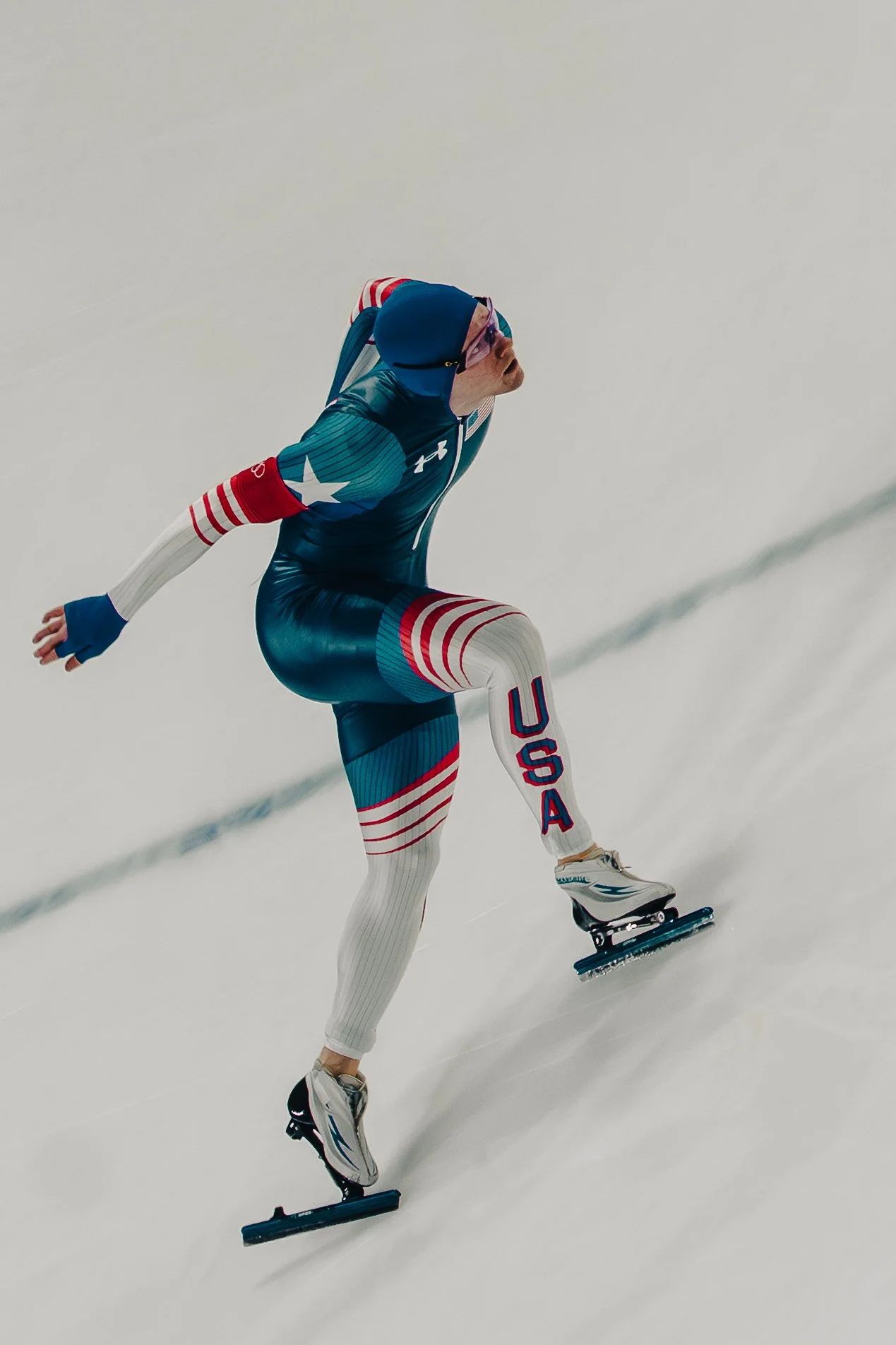 A speed skater dressed in a blue USA-themed suit with red and white striped accents, wearing glasses, skating on ice with arms extended and slightly bent, in a racing stance.