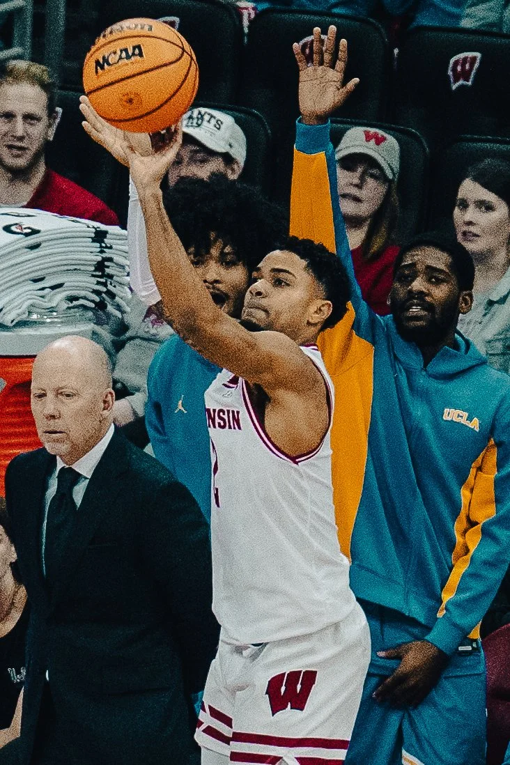 A basketball player in a white jersey from Wisconsin is taking a shot, with a crowd of spectators behind him. The crowd includes people wearing sports apparel and hats, with some raising their hands.