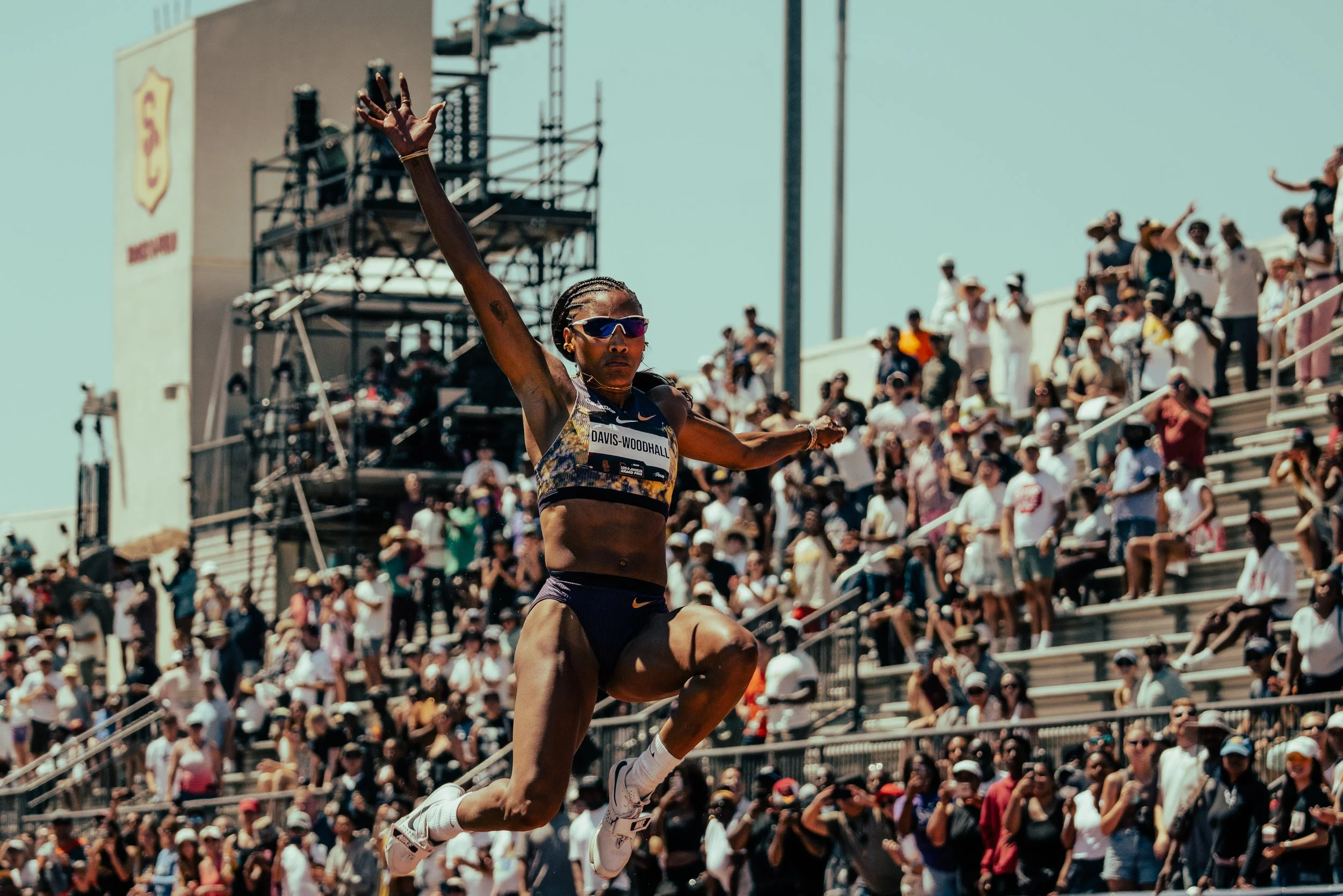 A female runner jumping at a track and field event with a large crowd in the stands behind her.