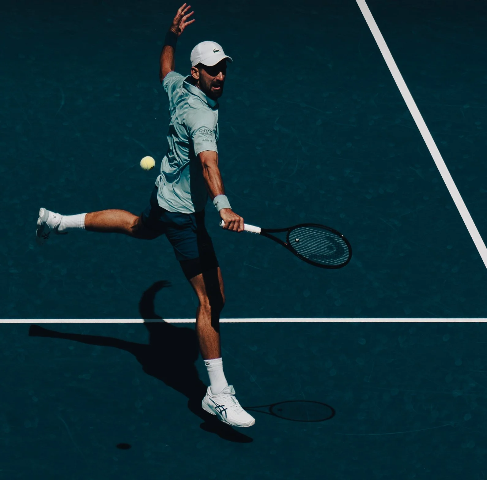 A male tennis player in a light blue shirt, black shorts, white cap, and white sneakers playing tennis on a blue court, hitting a tennis ball with his racket.