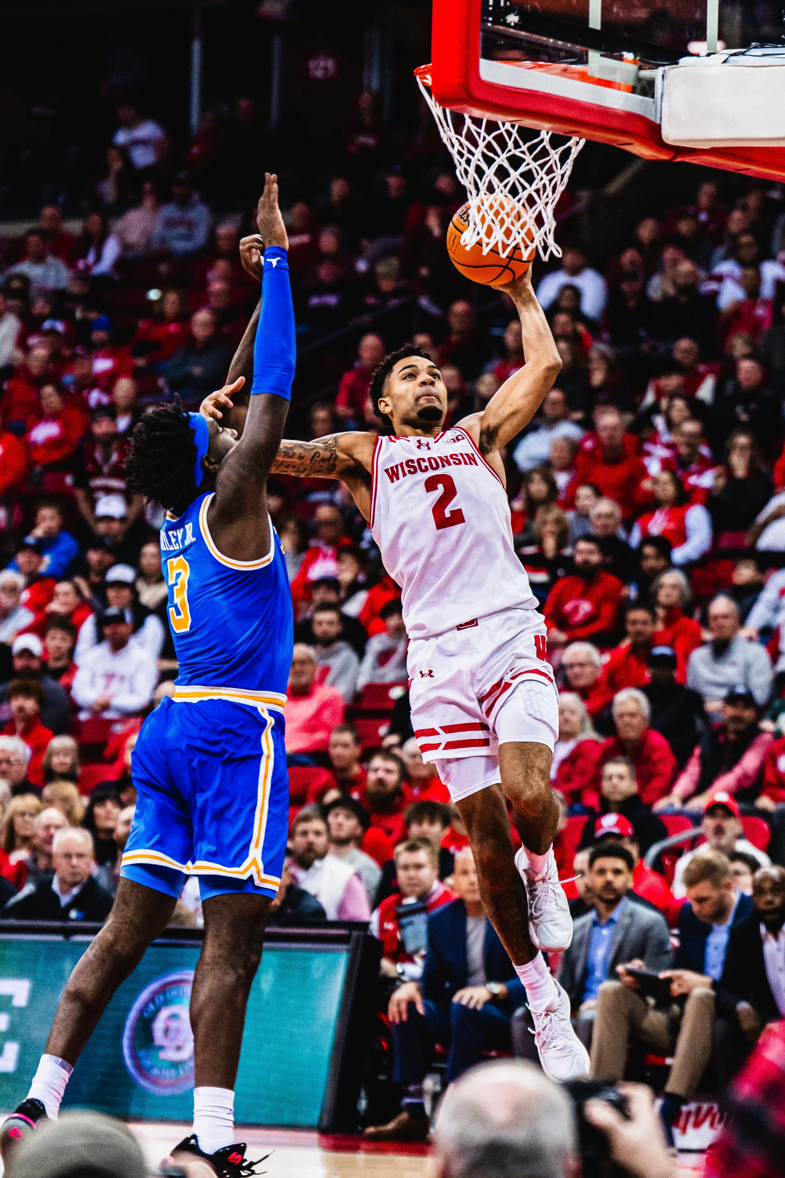 A basketball player wearing a Wisconsin uniform with the number 2 is jumping to score a basket while another player with a blue uniform and the number 3 attempts to block the shot during a game in an indoor basketball arena filled with spectators.
