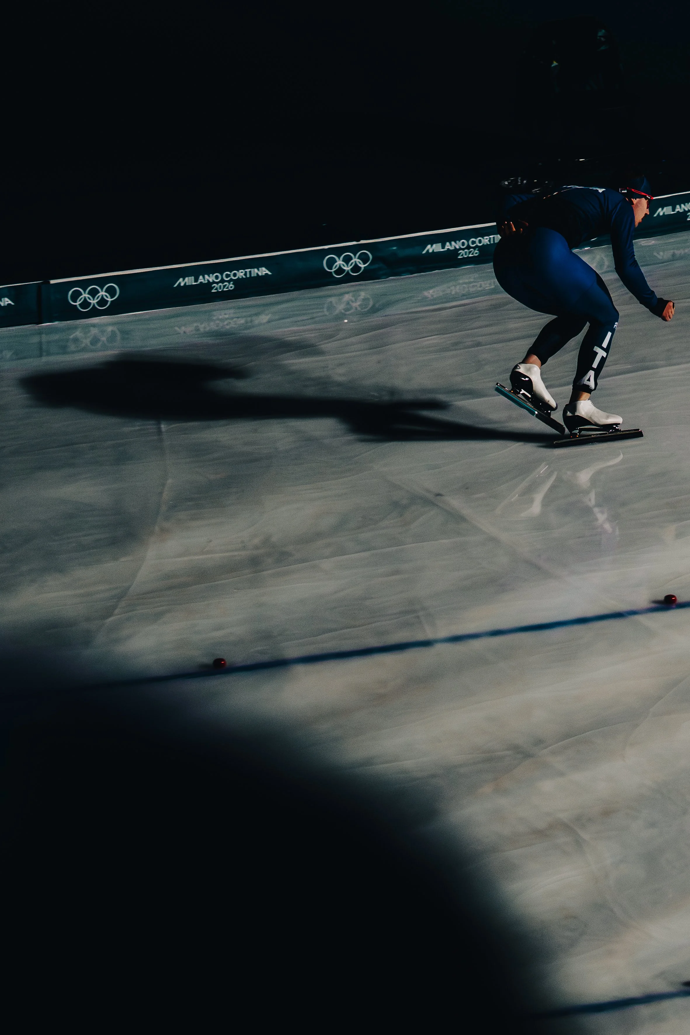 An athlete in athletic gear is ice skating at night during the Milano Cortina 2026 Winter Olympics, with a backdrop of dark ice and a display bearing the event's logo and Olympic rings.