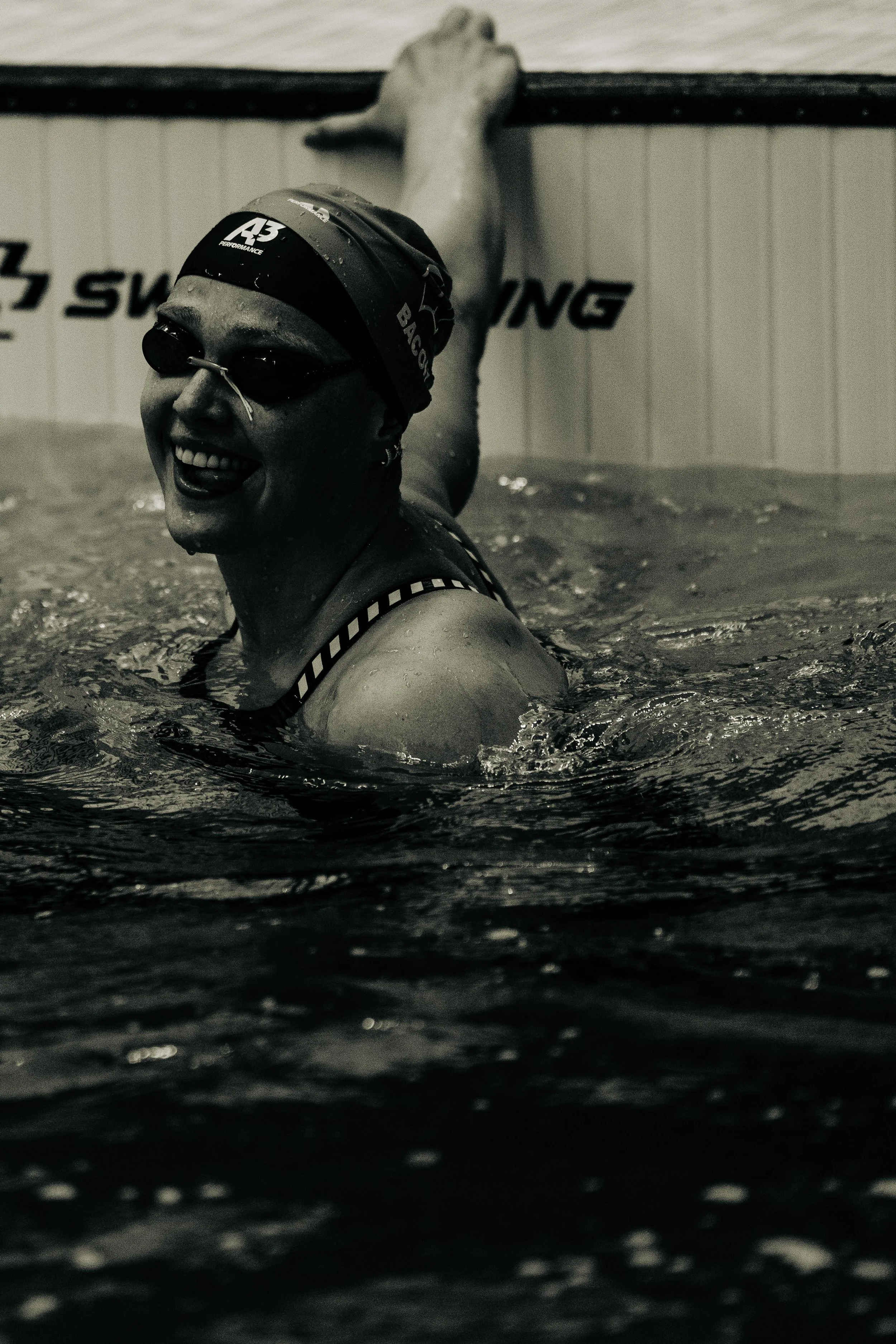 Swimmer smiling in an indoor swimming pool, wearing a swim cap and goggles, with water droplets on her face and swimming lane markings visible.