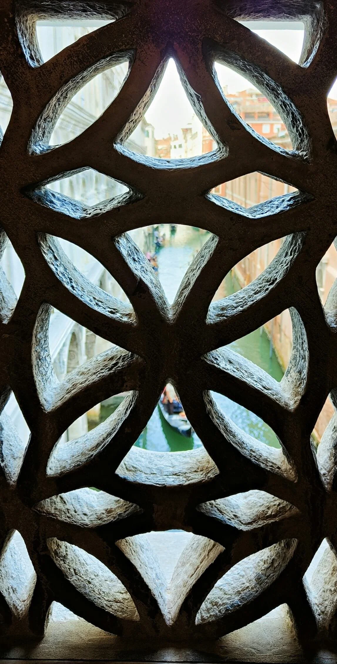Close-up view of an ornate, lattice-style, stone window or decorative panel with geometric patterns, with a canal and boat visible through the openings.
