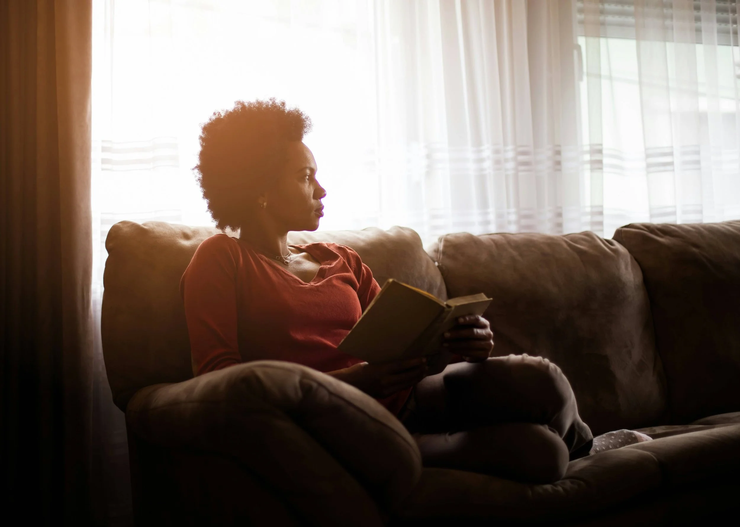 A woman with curly hair sitting on a couch, reading a book, with sunlight coming through sheer curtains in the background.