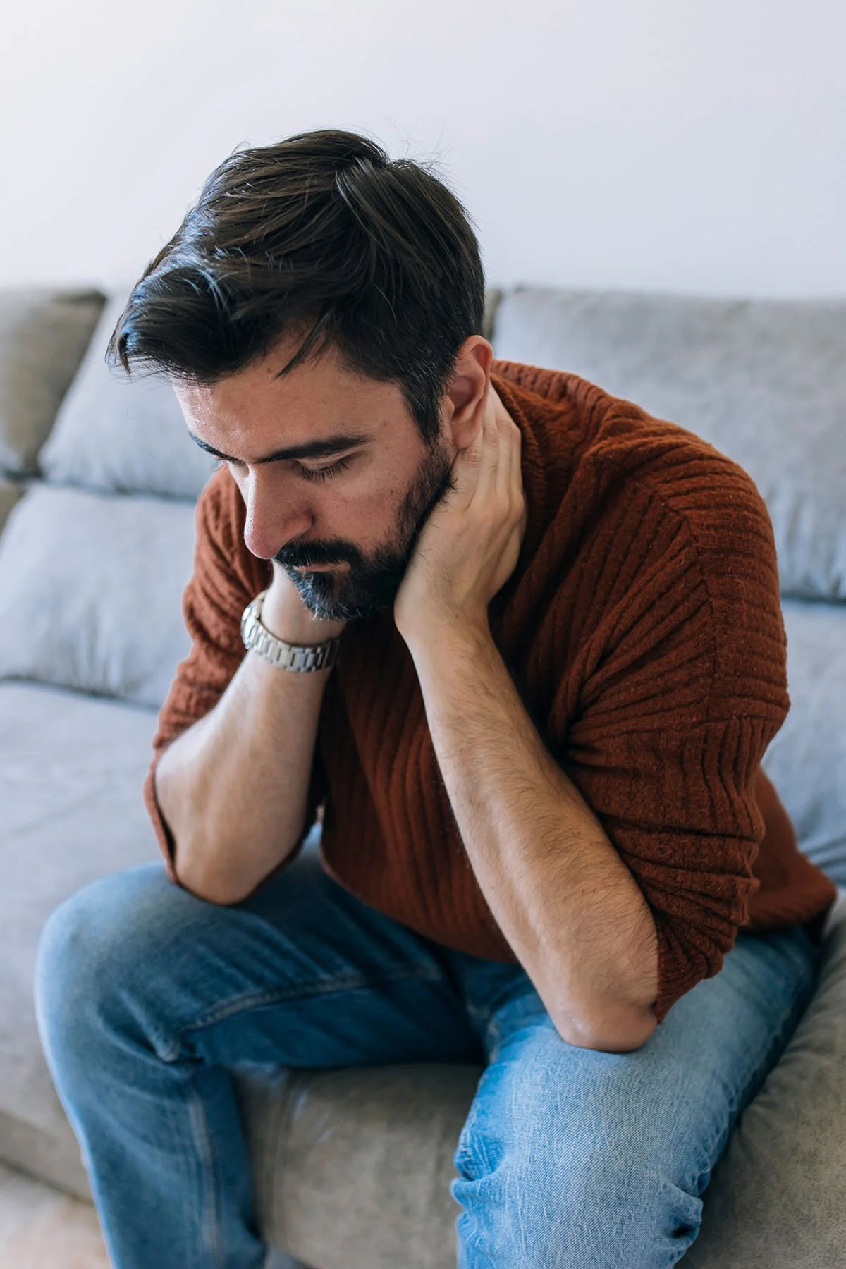 Man with dark hair and beard sitting on a gray sofa, holding his neck in pain, wearing a brown long-sleeve shirt and blue jeans.