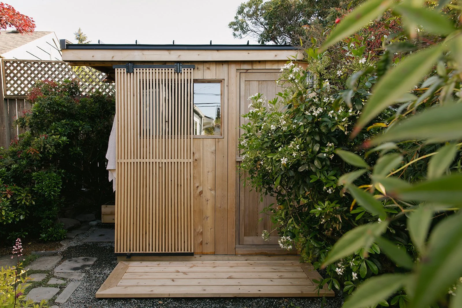 Small wooden sauna with a sliding door and a small window, surrounded by green bushes and plants, with a wooden deck in front. Seattle backyard sauna.