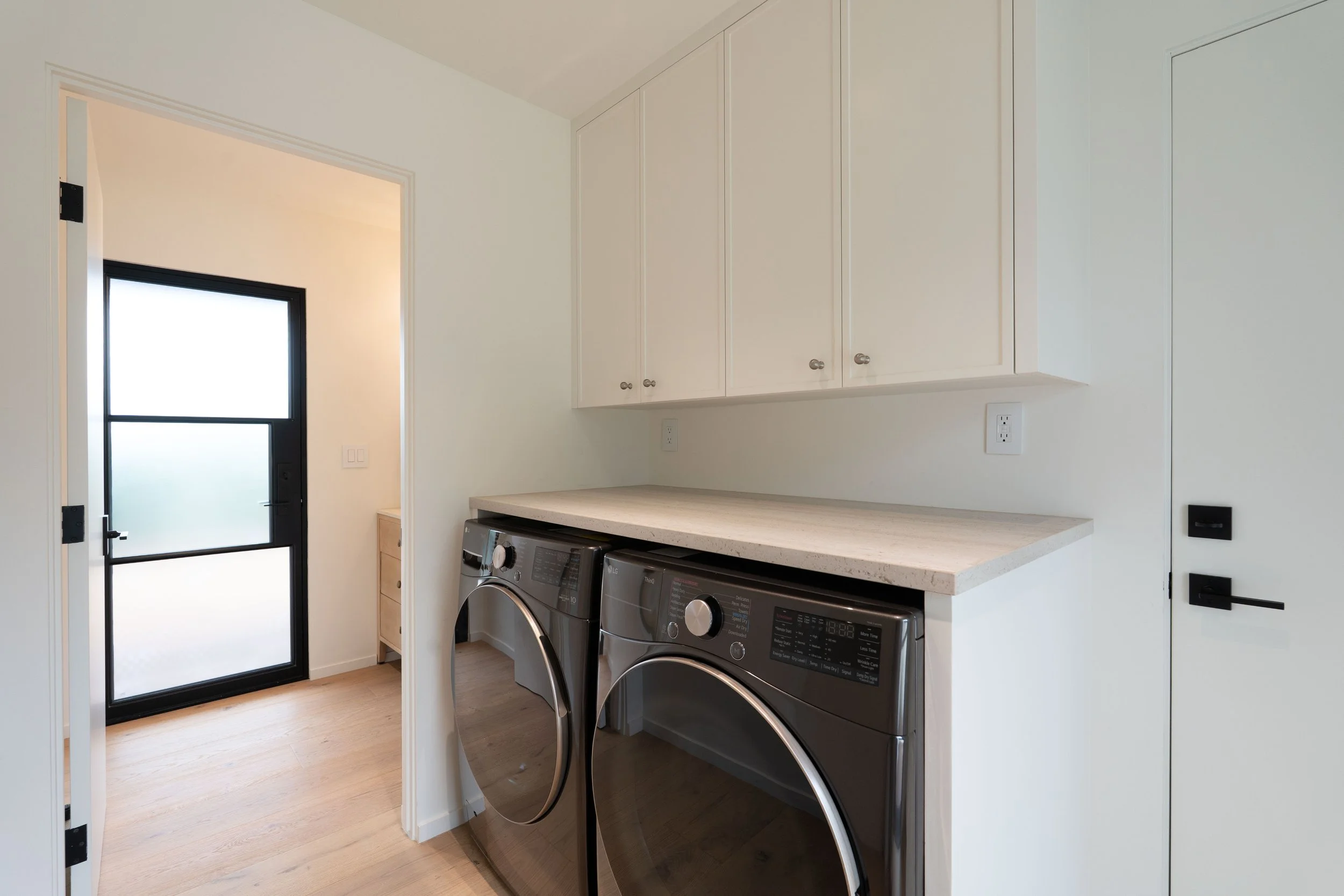 A laundry room with a white cabinet, washer and dryer, countertop, and a black-framed glass door.