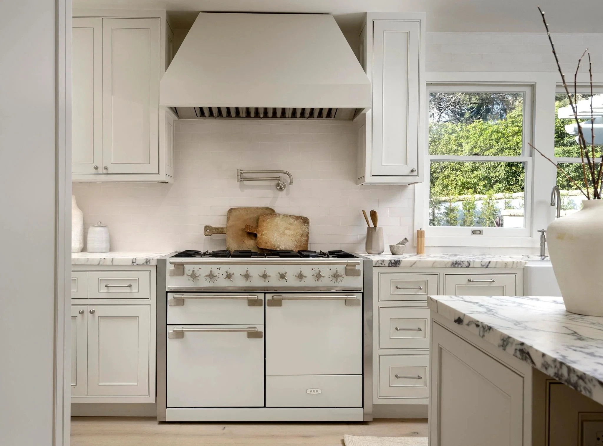 Modern white kitchen with marble countertops, white cabinets, a gas stove, and a large window showing greenery outside.