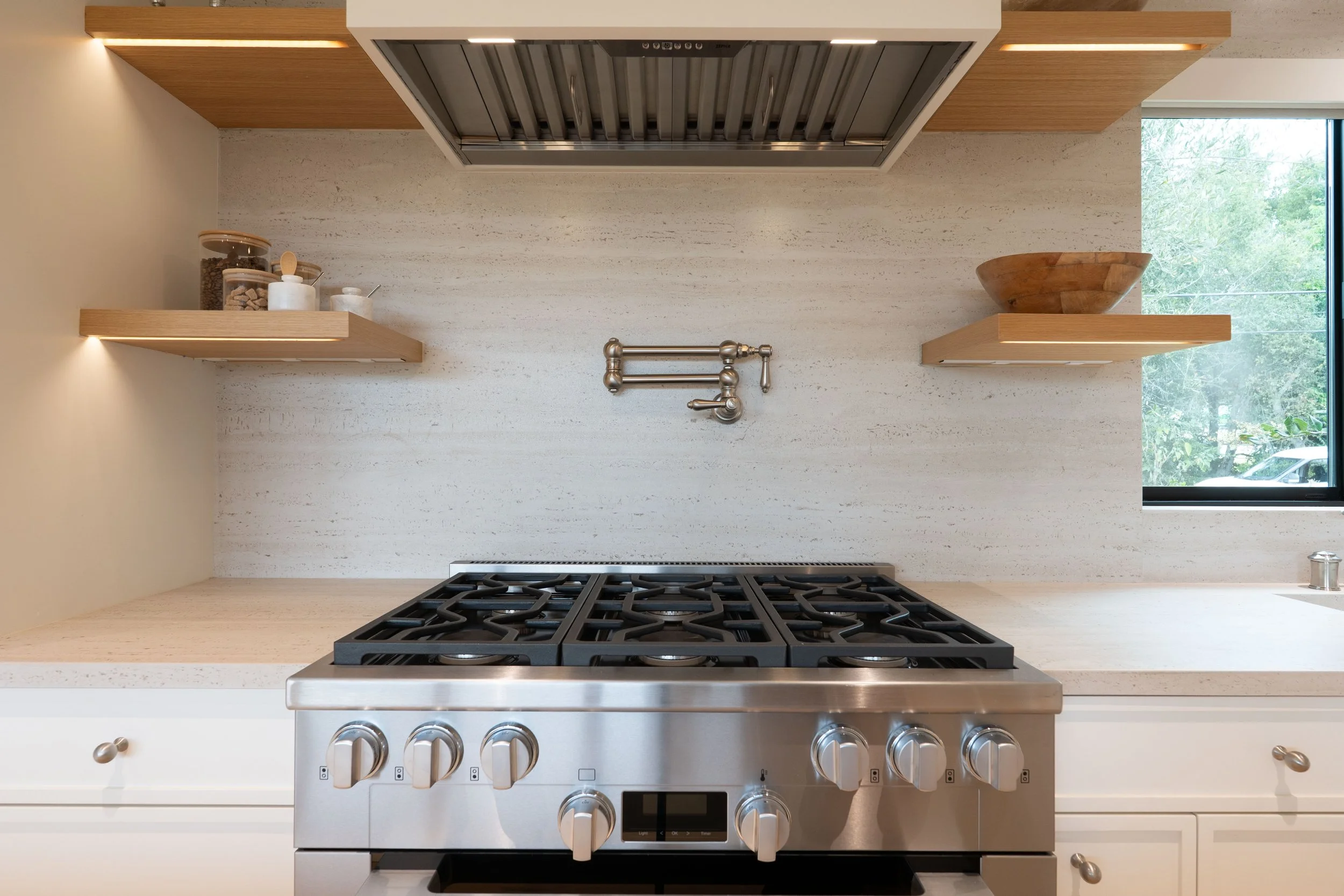 Modern kitchen stove with four burners, stainless steel finish, and digital display, positioned below a wall-mounted metal pot filler faucet.