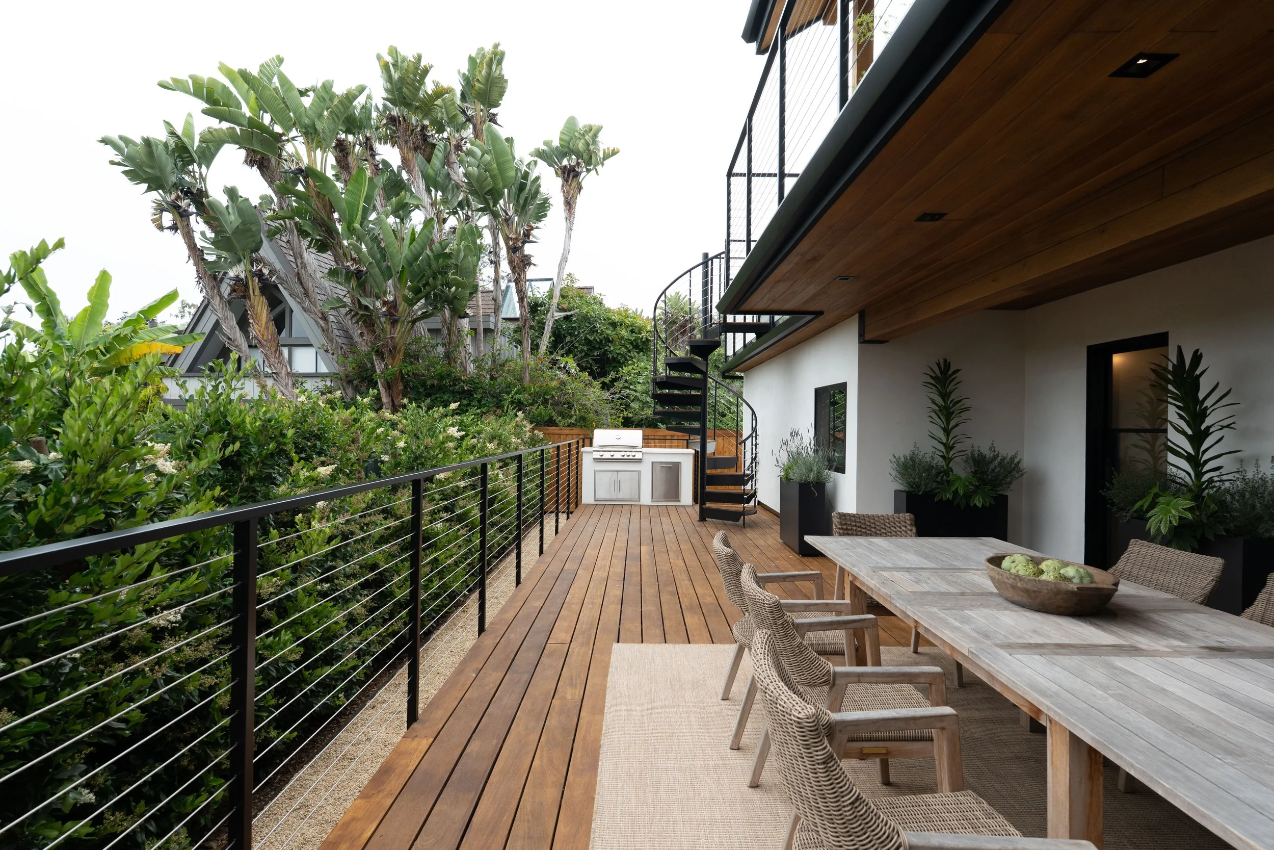 An outdoor patio with a wooden deck, surrounded by lush green plants and trees, featuring a dining table with chairs and a BBQ grill in a modern residential setting.