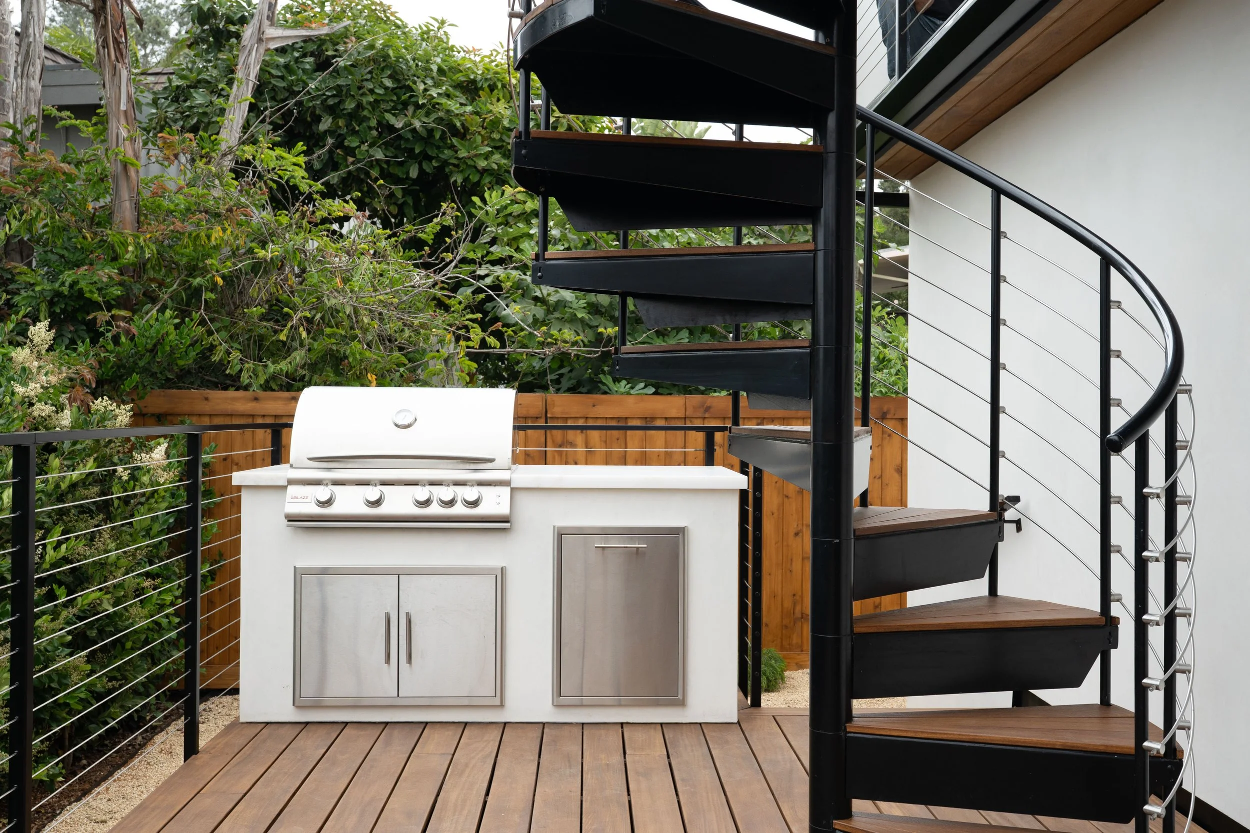 Outdoor balcony with a spiral staircase, BBQ grill, and outdoor kitchen setup, surrounded by greenery and wooden fencing.