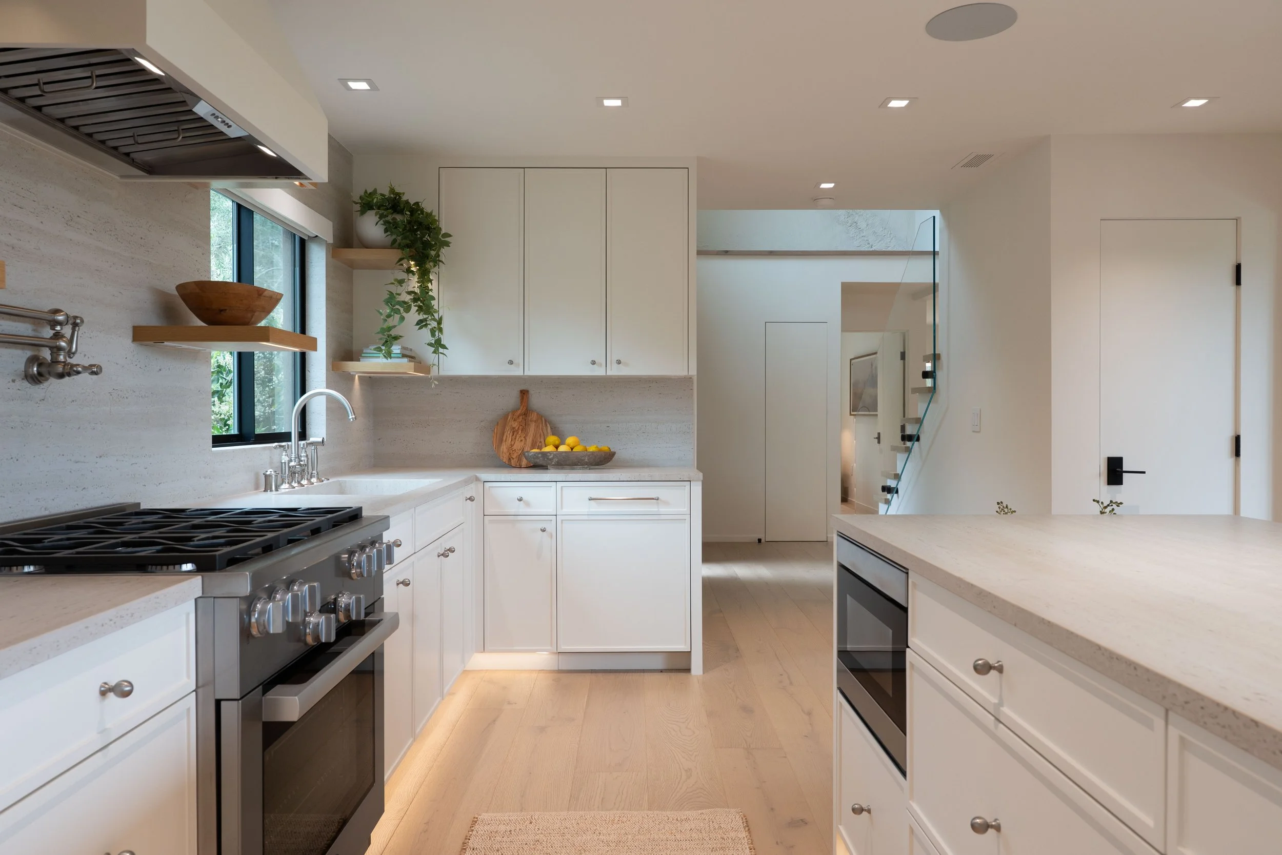Modern kitchen with white cabinets, beige countertops, and wooden flooring. Includes a stainless steel oven, a window with a view of greenery, and decorative elements like potted plants and a bowl of lemons.