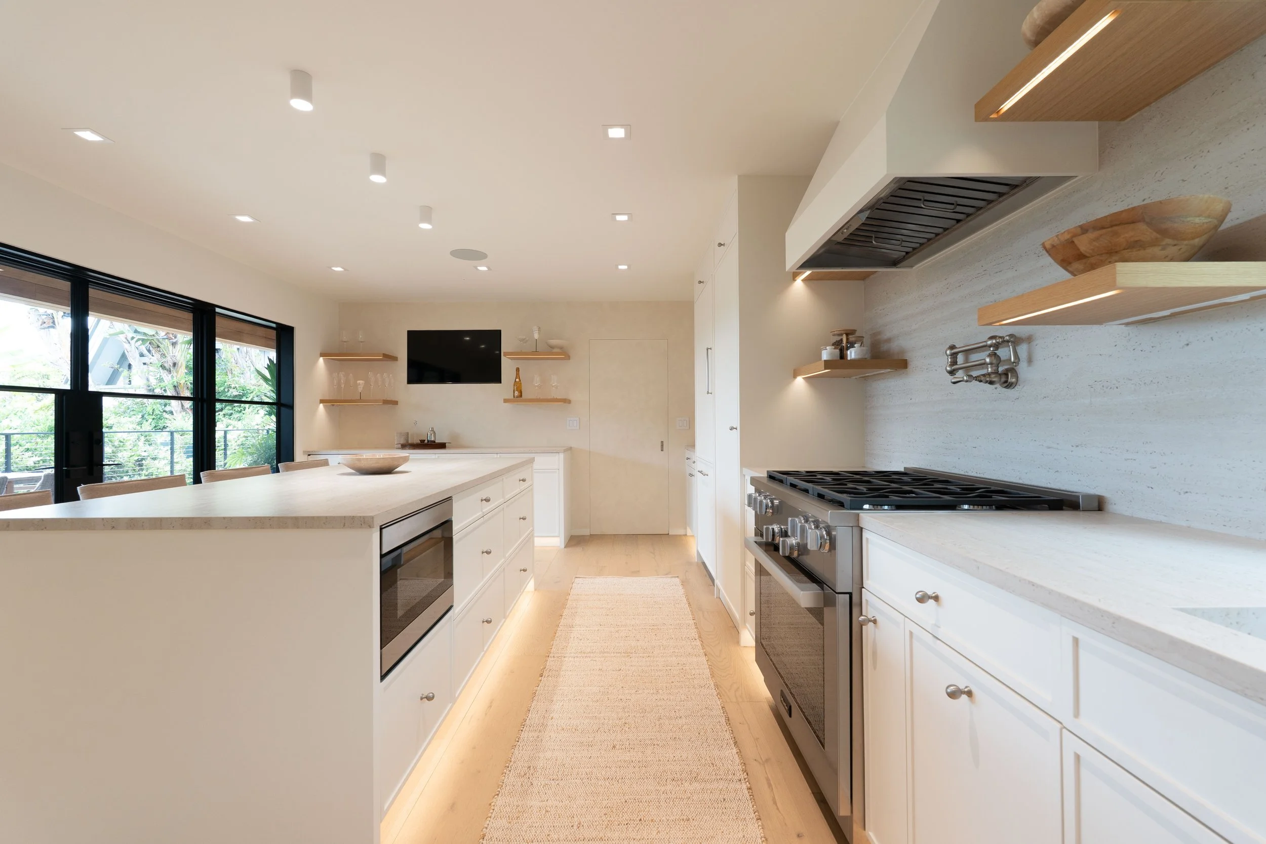 Modern kitchen with white cabinets, beige countertops, and built-in stainless steel appliances, including a stove. Open shelves hold glassware and wooden bowls. Large sliding glass doors lead to an outdoor balcony with greenery.