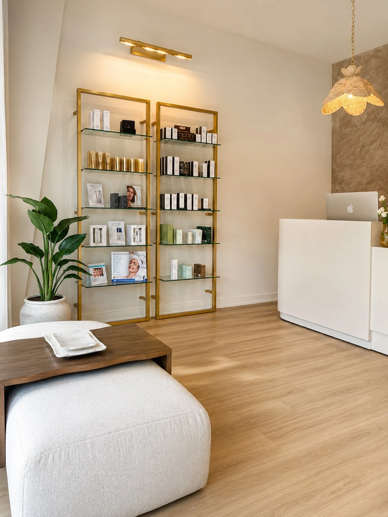 A modern, minimalistic reception area with a white curved reception desk, a small wooden table with white towels, a large potted green plant, and two gold metal and glass display shelves with skincare or beauty products. There is a partial view of a white upholstered armchair in the foreground, wood flooring, and a textured beige wall. A decorative hanging lamp and a wall-mounted gold light fixture illuminate the space.