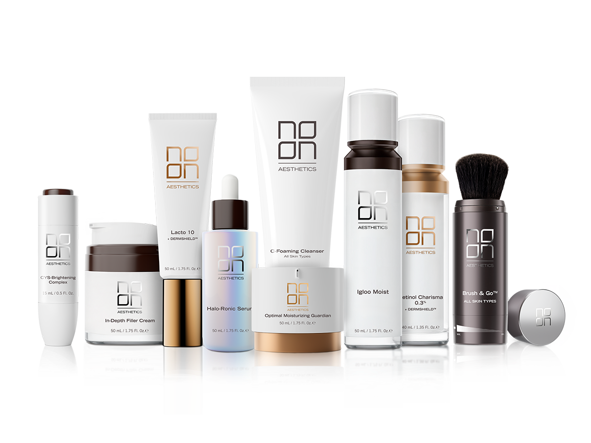 A collection of skincare and cosmetic products in white, black, and gold packaging, including creams, serums, cleansers, sprays, a brush, and a jar, arranged on a reflective surface with a black and white background.