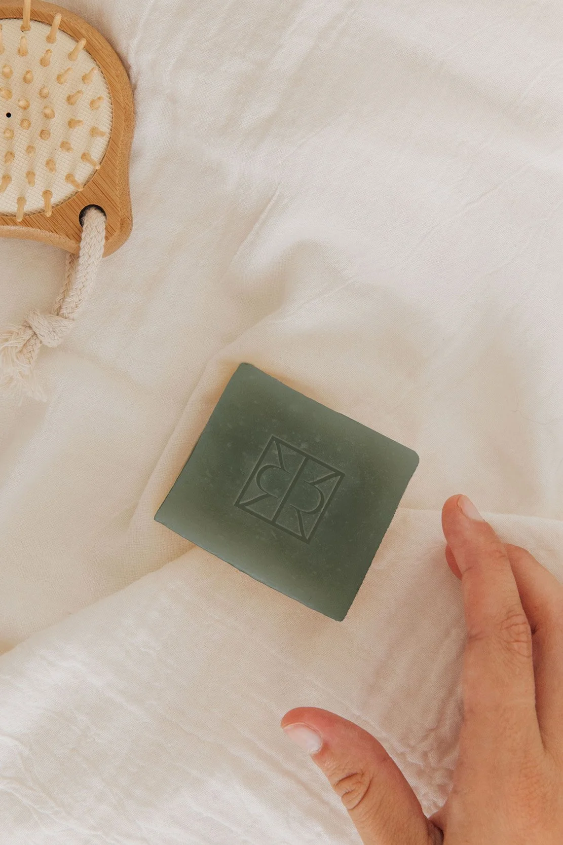 A hand holding a green soap or stone with a carved The Row logo on it, placed on a white fabric surface with part of a wooden spa brush visible in the background.
