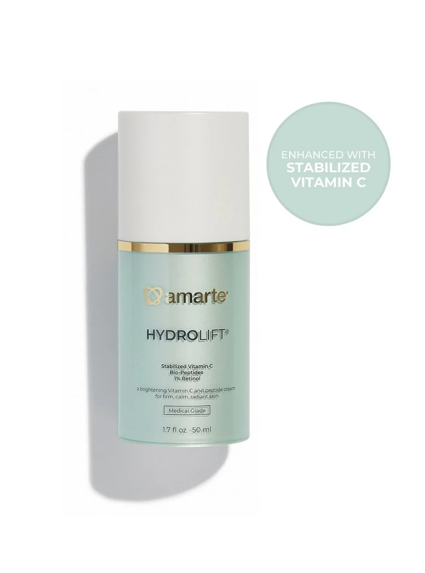 A white bottle of Amarte HydroLift moisturizer with a gold and white cap, placed on a white surface. There is a light green circular label on the right side of the bottle indicating it is enhanced with stabilized vitamin C.
