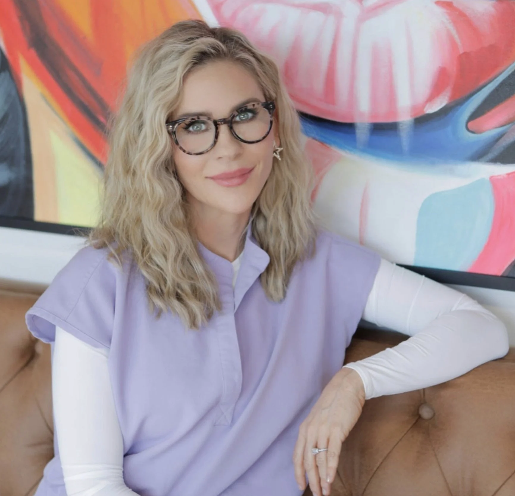A woman with blonde wavy hair, wearing glasses and a lavender medical smock, sitting at a wooden table with her hands resting on her lap. Behind her is colorful abstract artwork on the wall.